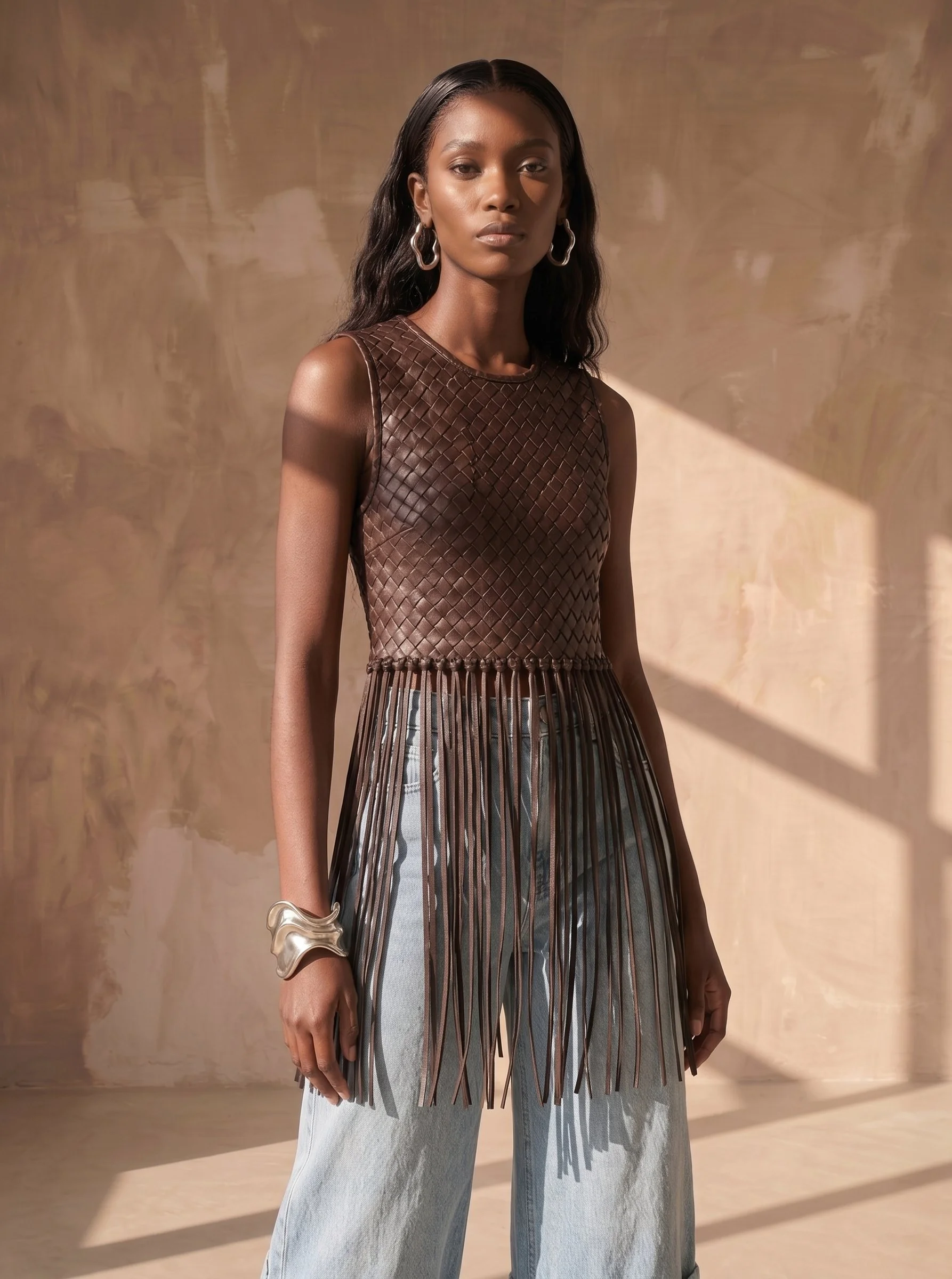 A woman with dark skin and long black wavy hair wears a sleeveless brown leather top with a woven pattern, large silver hoop earrings, a silver bracelet, and light blue wide-legged jeans.