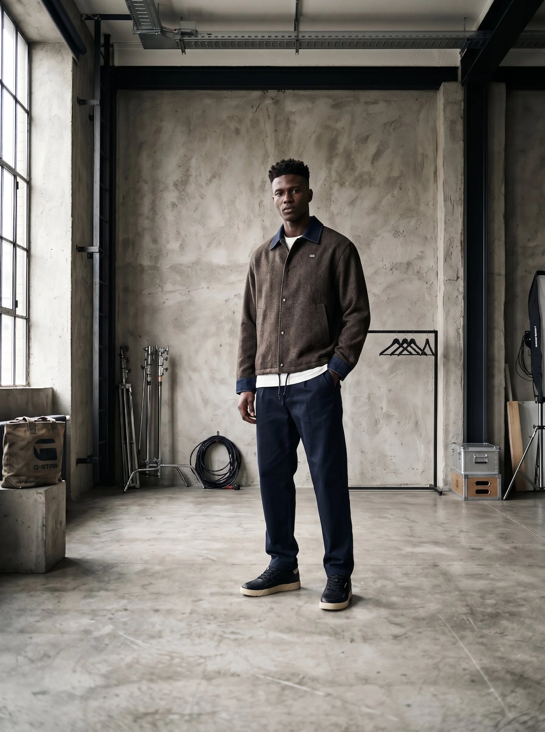 A young man standing in an industrial-style room with concrete walls, metal pipes, and a large window, wearing a brown jacket, dark pants, and black sneakers.