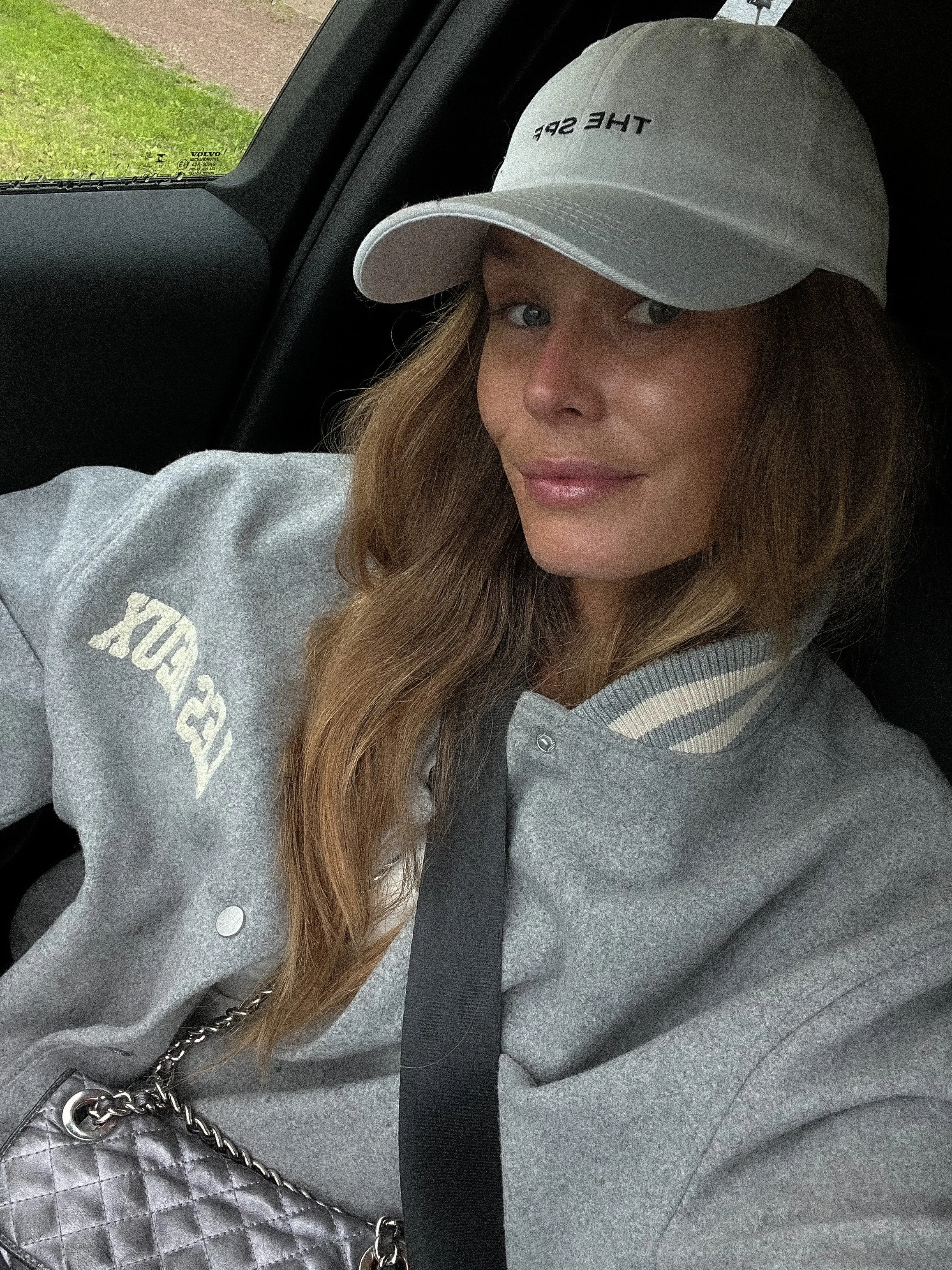 A woman with long light brown hair wearing a light gray hoodie with white and gray striped cuffs, a white baseball cap that says "THE SP" on it, and a silver quilted purse with a chain strap, sitting in a vehicle.