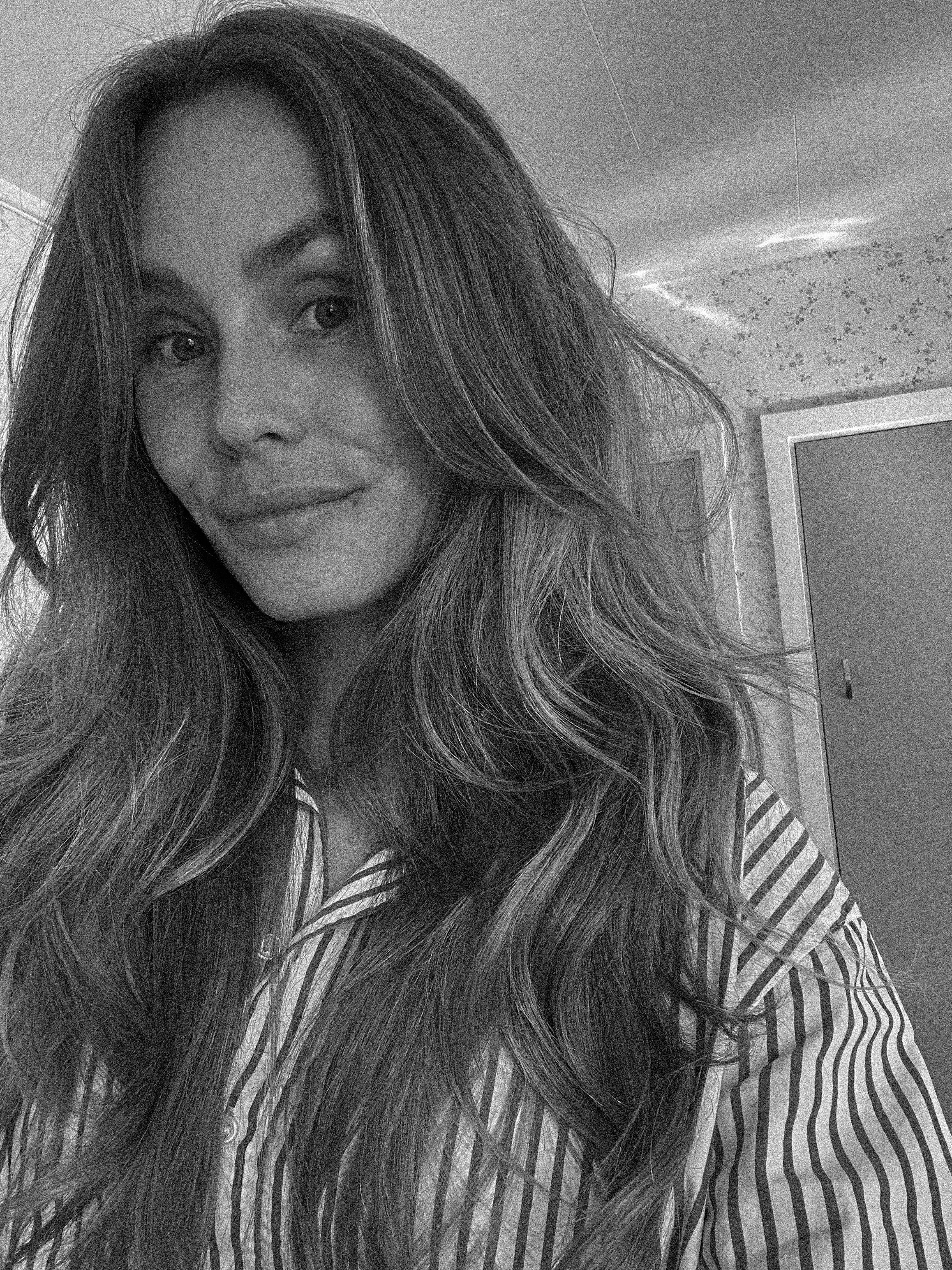 Close-up black-and-white photo of a woman with long wavy hair, smiling slightly, wearing a striped shirt, in an indoor setting with a textured wall and a TV in the background.