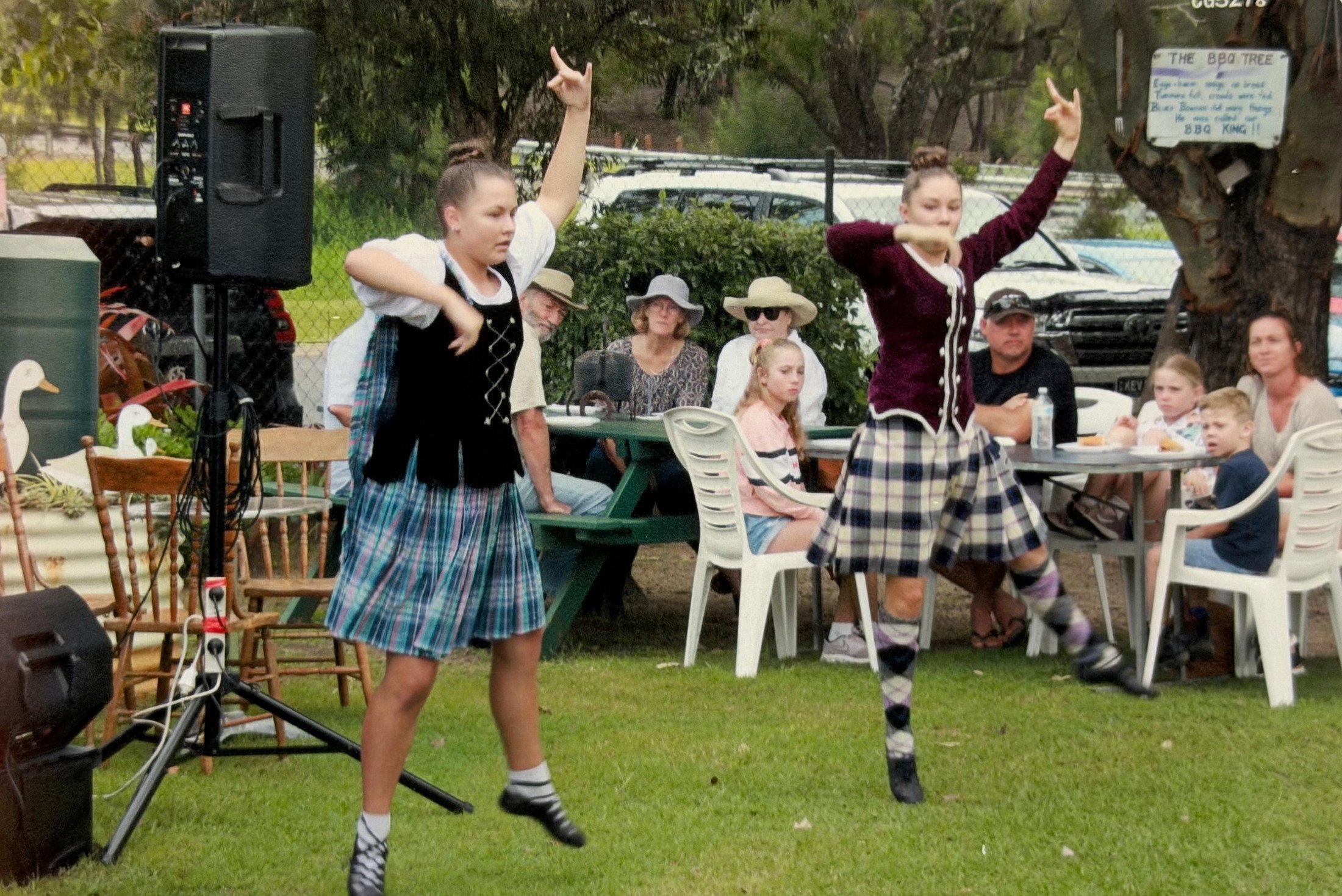 Two women dressed in traditional Scottish kilts and white socks are performing a Highland dance outdoors. Several onlookers, including children and adults, are watching them. The background features trees, parked cars, and a sign about a barbecue tree.
