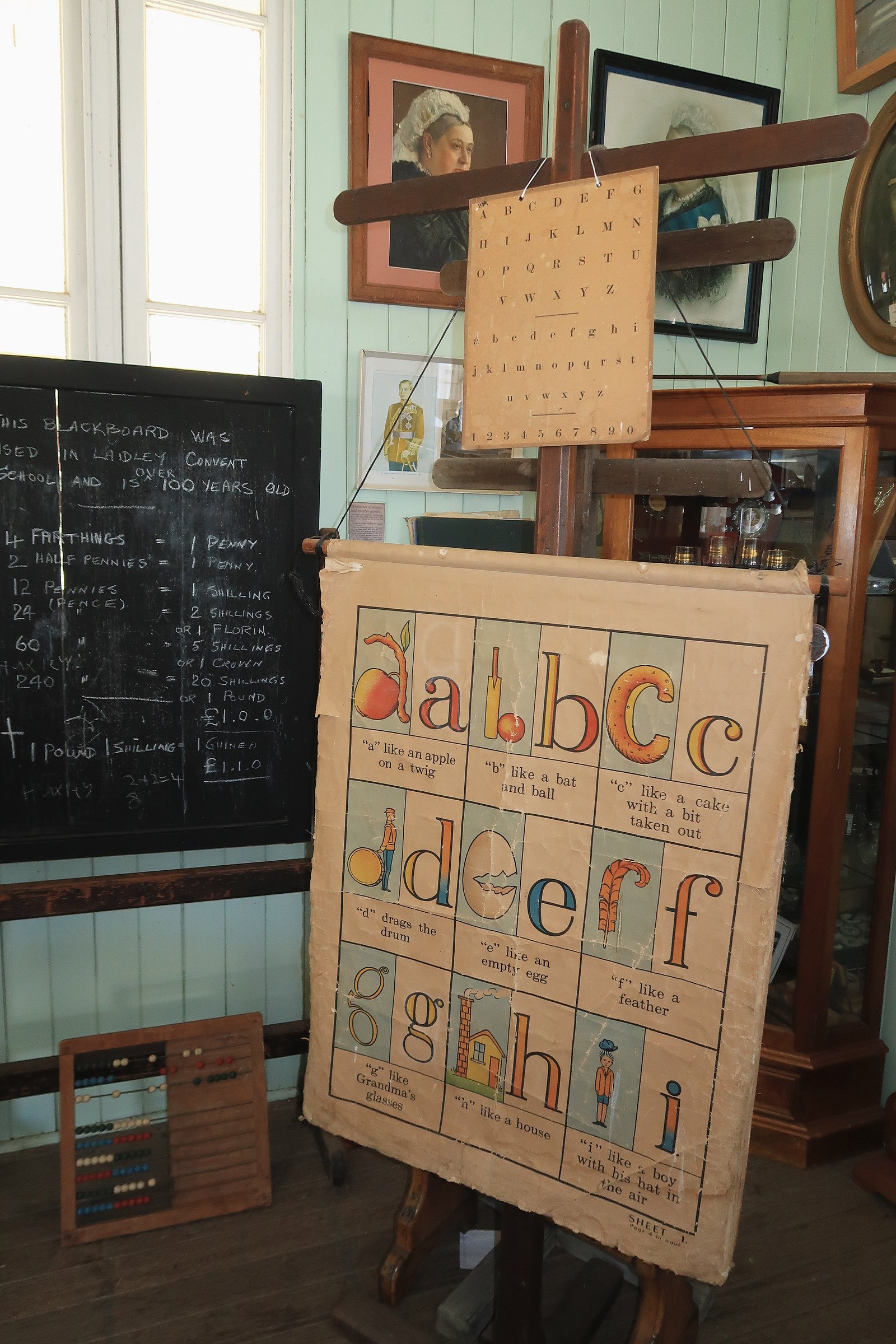 Educational display with vintage-style alphabet poster illustrating words for each letter, a blackboard with handwritten notes, and traditional toys, all inside a room with framed artwork and wooden furniture.
