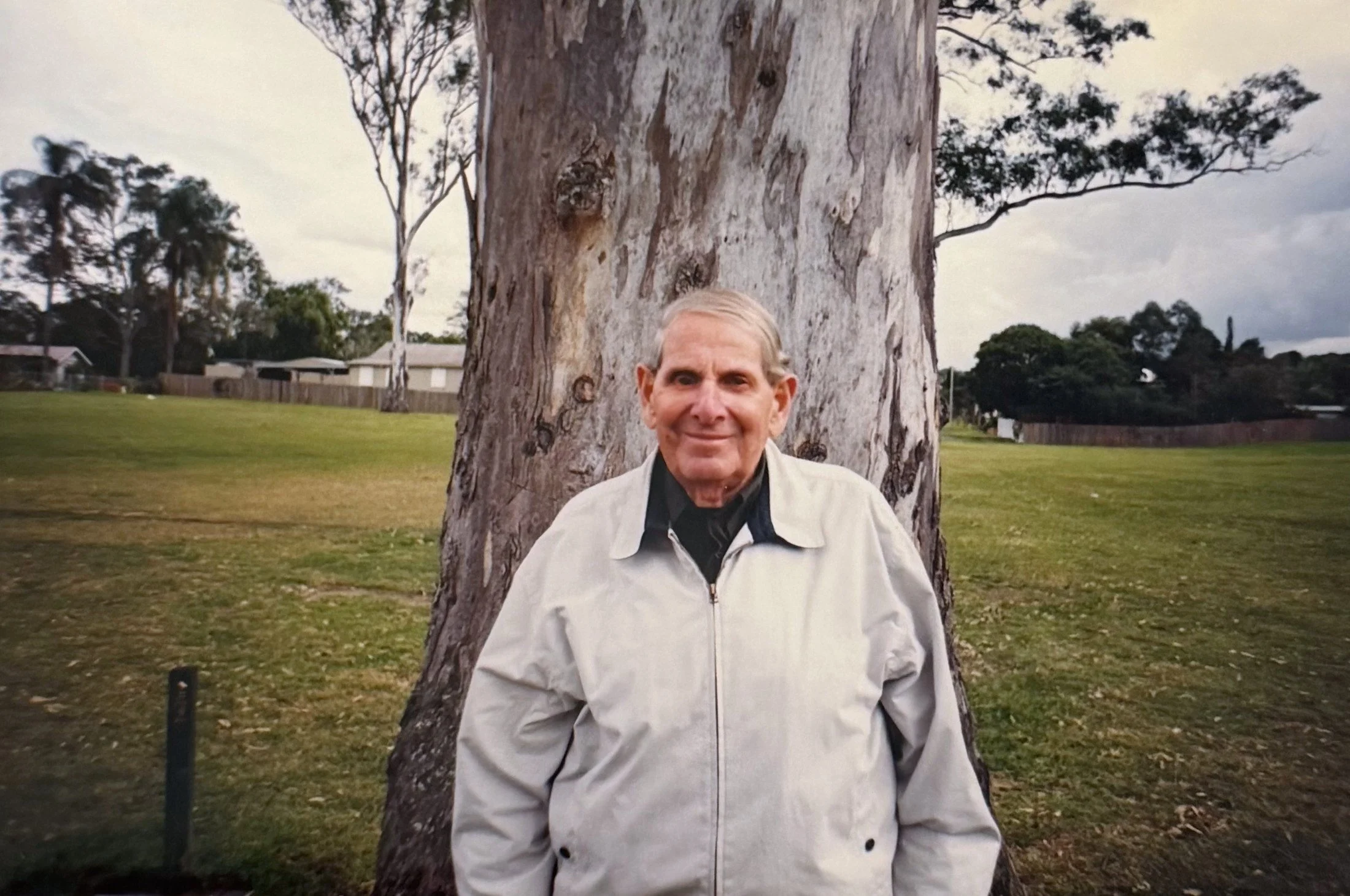 An elderly man with gray hair smiling, standing in front of a large tree in a grassy park with cloudy sky and residential houses in the background.