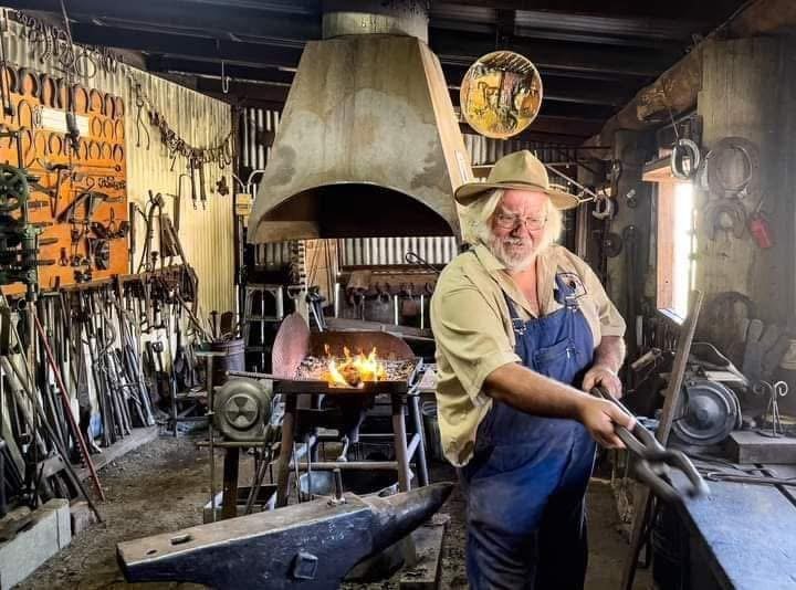 A blacksmith wearing a cowboy hat and apron working inside a blacksmith shop with tools hanging on the wall and a forge with flames behind him.