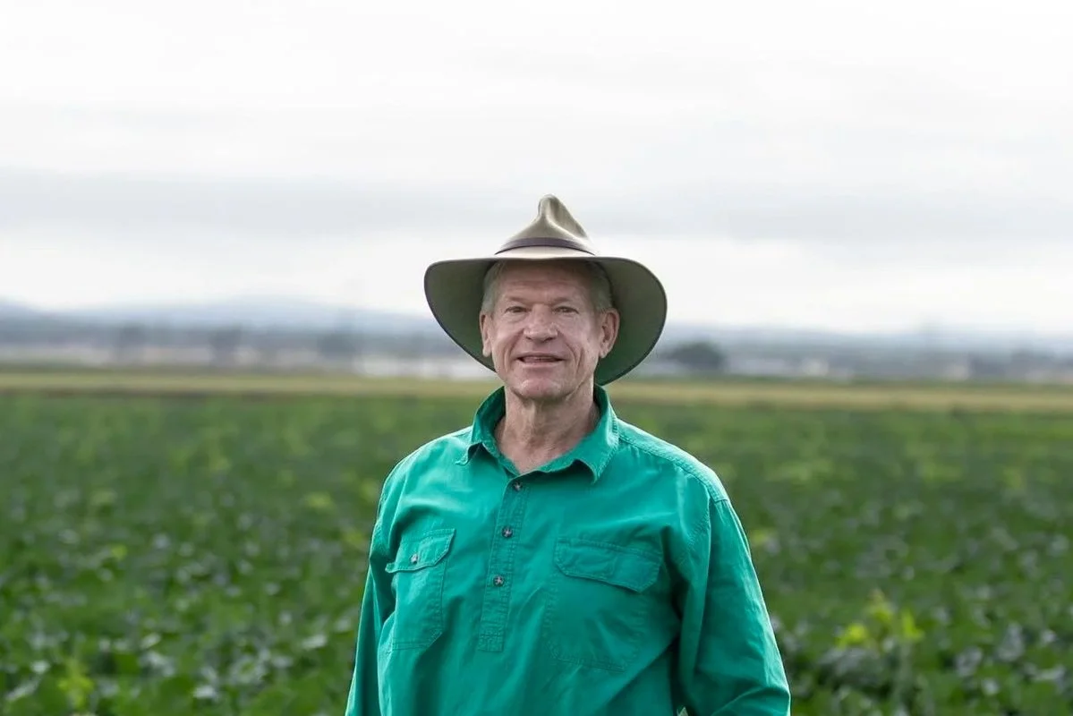 A man standing in an agricultural field wearing a wide-brimmed hat and a green button-up shirt.