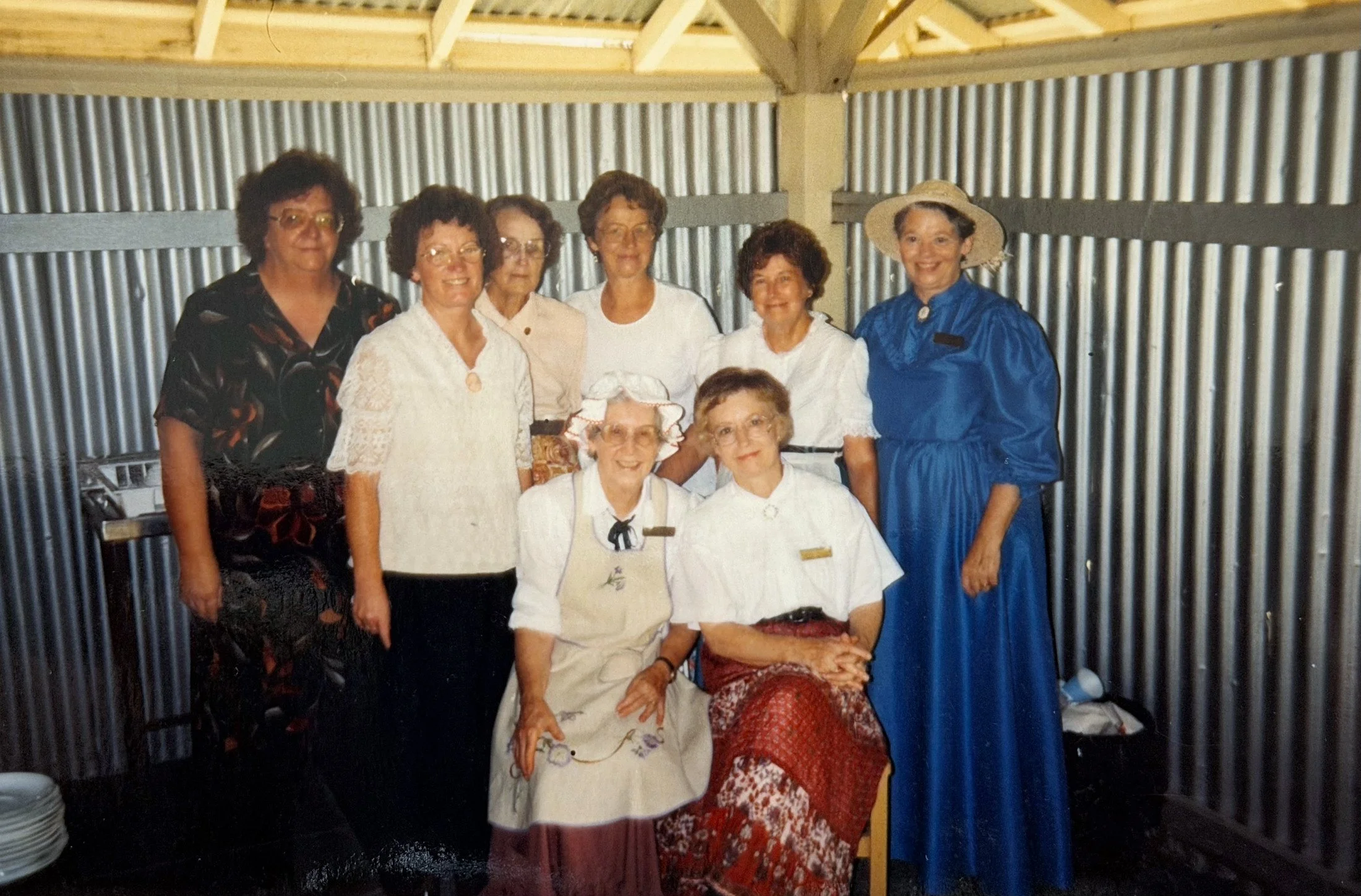 Group of women, mostly elderly, gathered together indoors, some wearing vintage or traditional clothing, posing for a photo with a wooden and corrugated metal wall in the background.