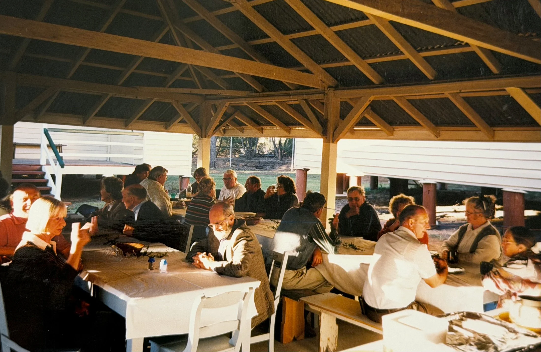 People sitting and talking at a long table in an outdoor patio area with a covered wooden roof, during what appears to be a social gathering or dinner event.