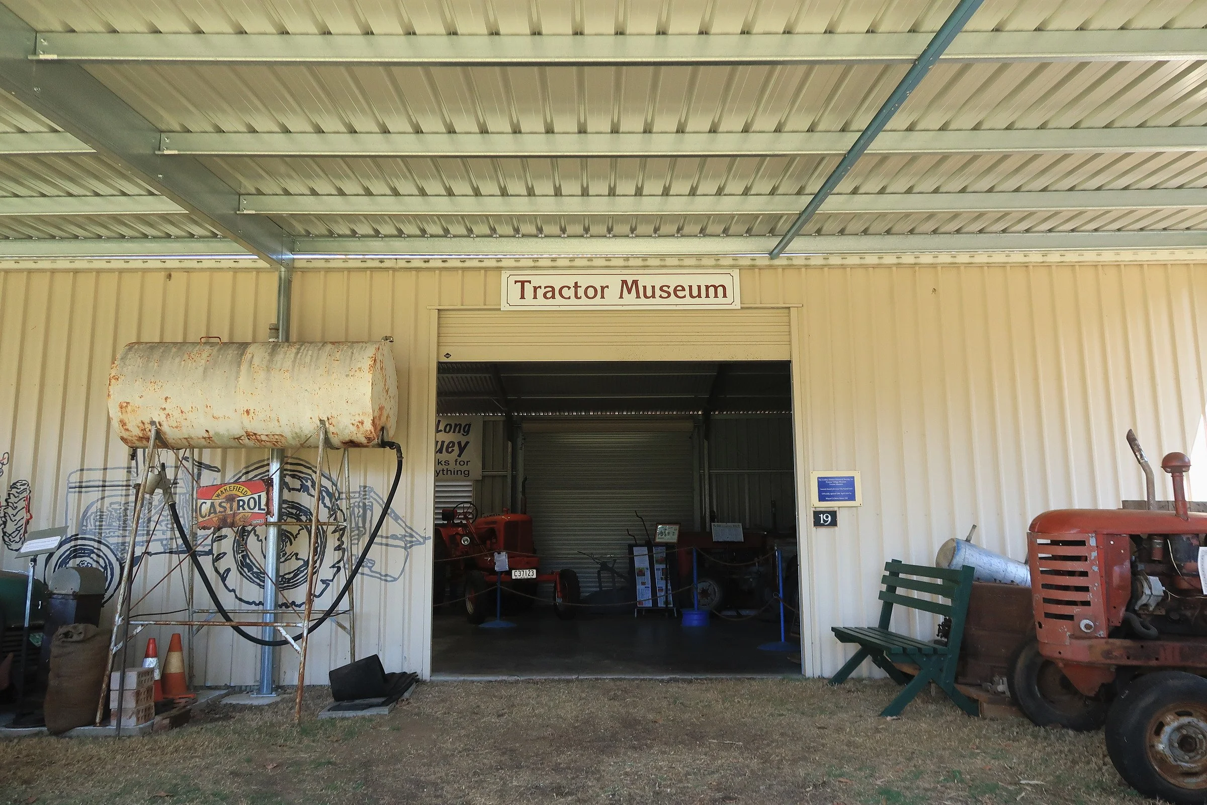 Entrance to the Tractor Museum with vintage farm equipment, including a rusty tank, model tractors, a bench, and various signs, all under a metal building with a sign reading 'Tractor Museum'.