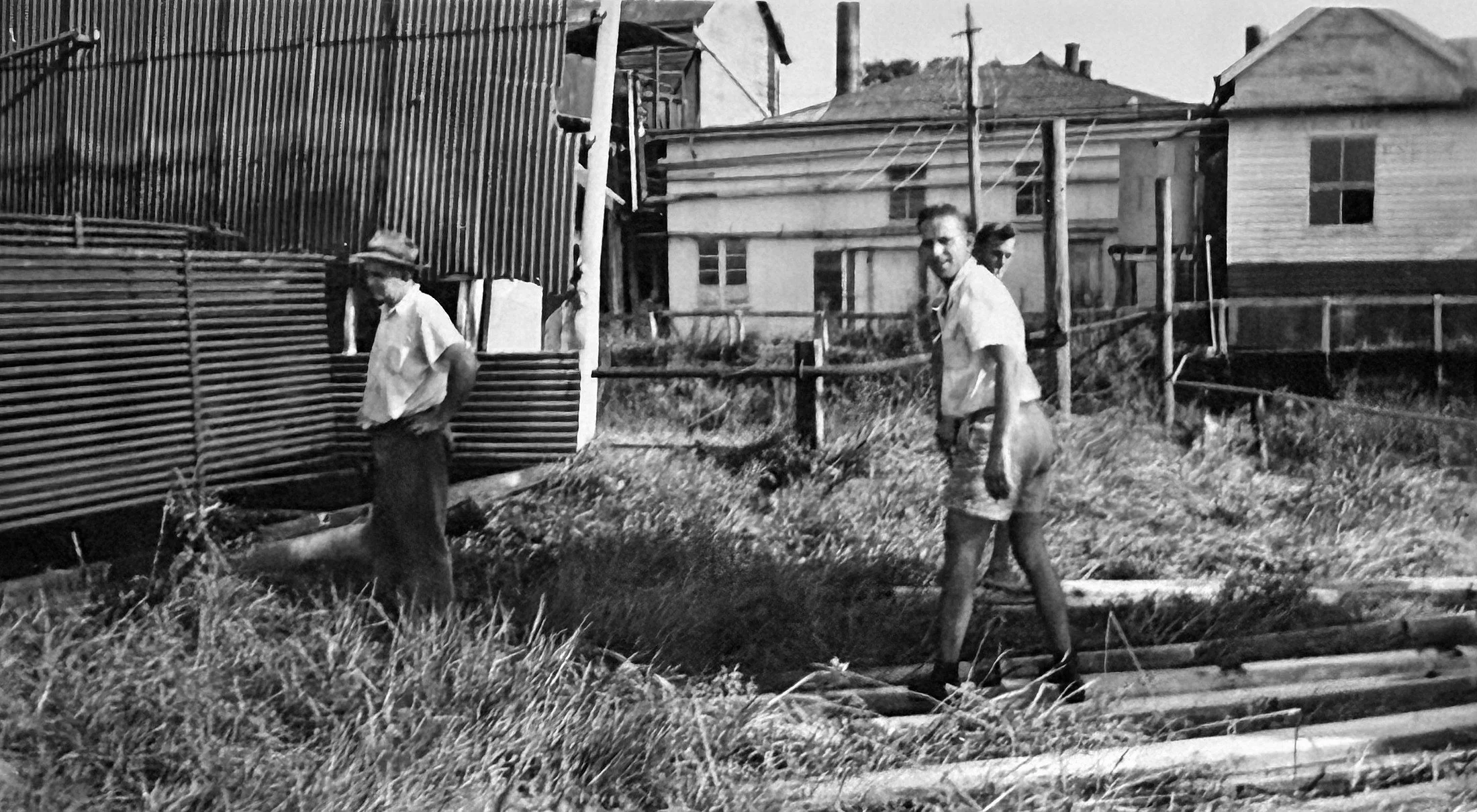 Two young boys walking outdoors in a residential area, one wearing a hat and the other with short hair, with houses and utility poles in the background.