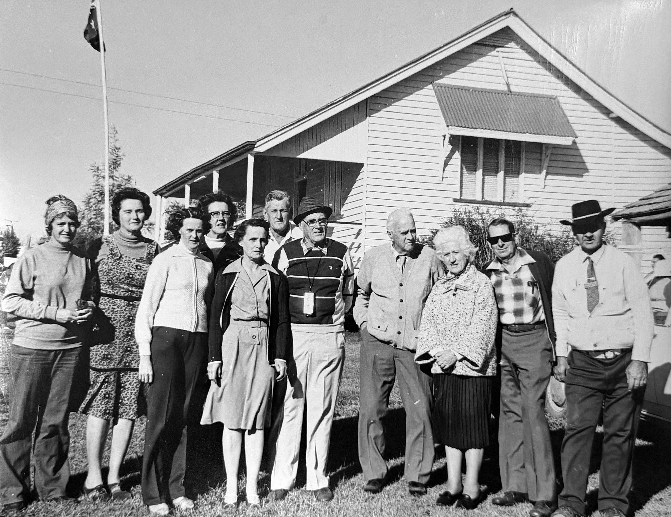 A black and white photo of a group of adults standing outside in front of a house with a flagpole. The group appears to be at a social gathering or event, with several women and men dressed in casual and semi-formal attire, some wearing glasses and hats.