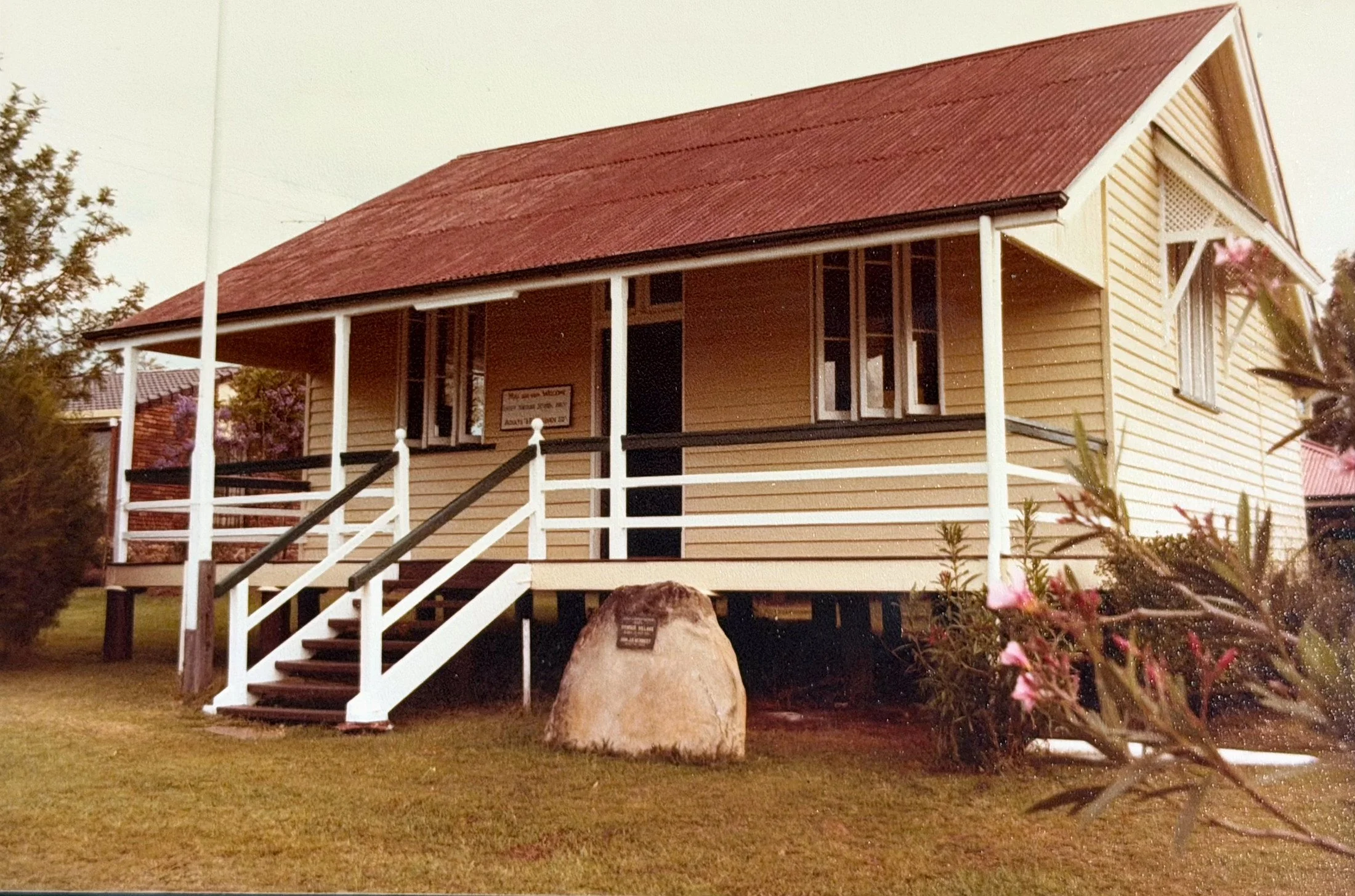 A yellow wooden house with a red roof, white trim, and a front porch with white stairs and railing, surrounded by grass and bushes.
