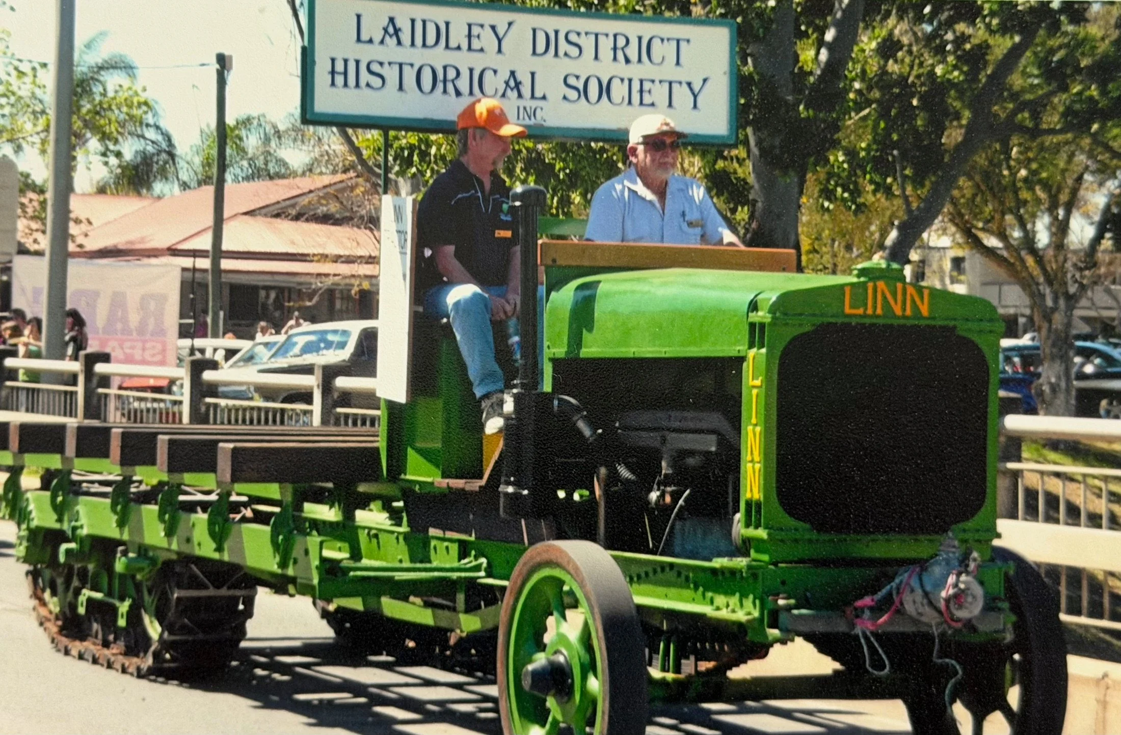 Two men riding on a bright green tractor with a sign above that reads 'Ladley District Historical Society Inc.' Trees and buildings are visible in the background and the men are wearing hats, one with an orange cap and the other with a tan cap.
