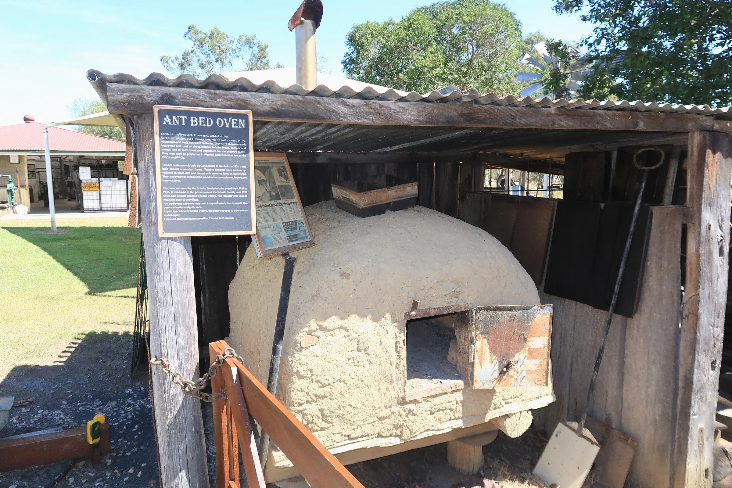 Historical outdoor oven constructed with clay and bricks, covered with a wooden roof and enclosed by wooden and brick walls in a park or museum setting.