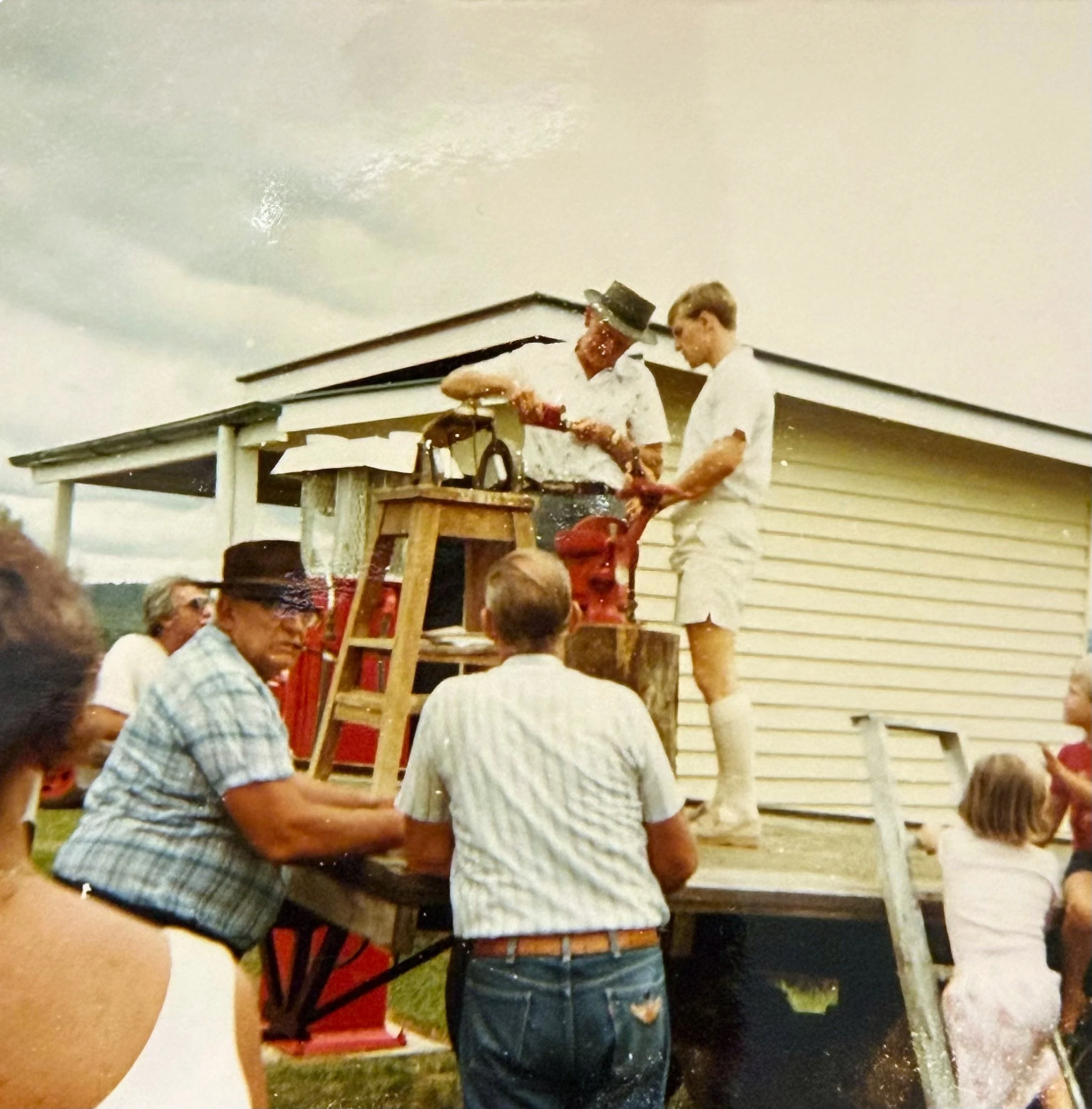 People gathered outdoors during daytime, with two individuals standing in front of a house, one in a white shirt and the other in a red shirt, engaging in an activity involving a red box or object. The background shows a house with horizontal siding and several other people, some seated and some standing.