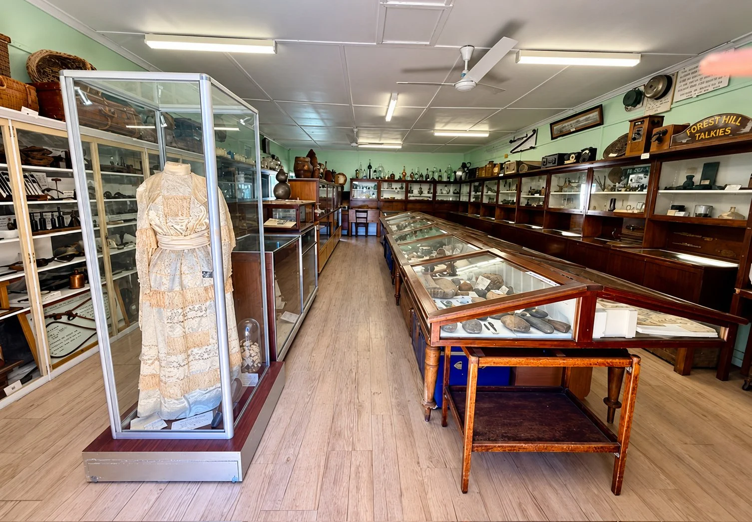 Interior of a museum or antique store with glass display cases filled with rocks and artifacts, wooden shelves and cabinets showcasing various collectibles, and a mannequin dressed in vintage clothing.