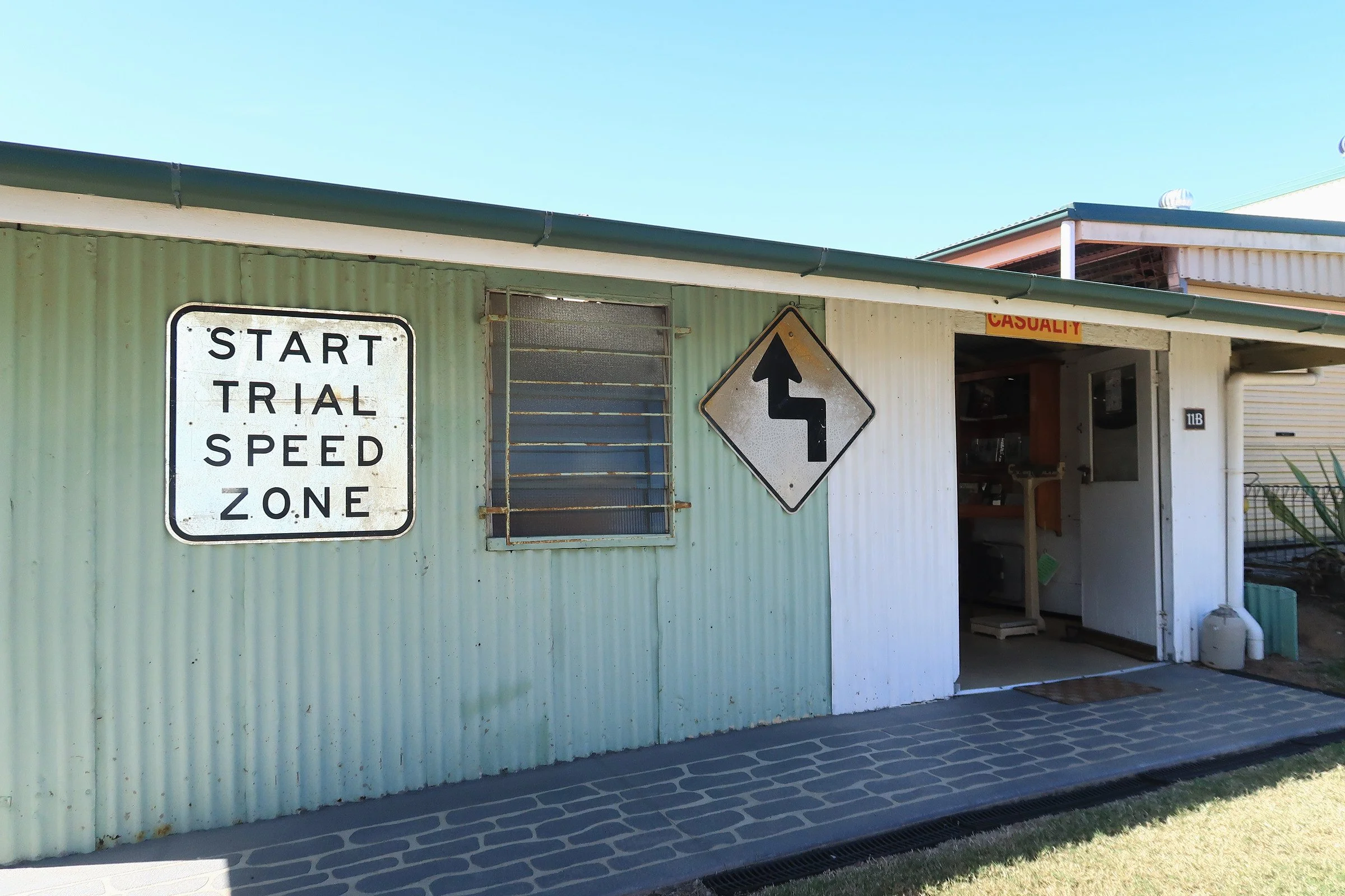 A building with a corrugated metal exterior painted light green, featuring a sign that reads 'START TRIAL SPEED ZONE,' a black arrow sign, and a small open doorway with the word 'CASUALTY' in red above it. The building is adjacent to a black, patterned drainage grate on the sidewalk, with a blue sky overhead.