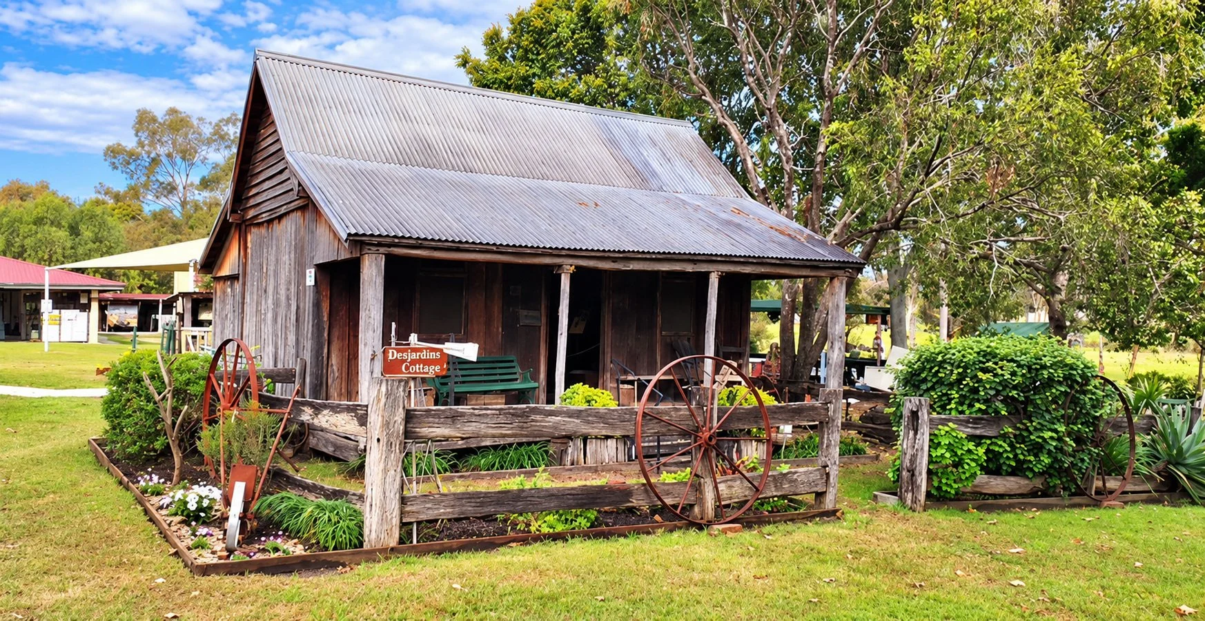 A rustic wooden cottage with a corrugated metal roof, surrounded by a garden with plants and flowers. There are old wagon wheels as decoration, a small sign reading 'Desjardins Cottage,' and a wooden fence. Trees and a building with a red roof are in the background on a bright, clear day.