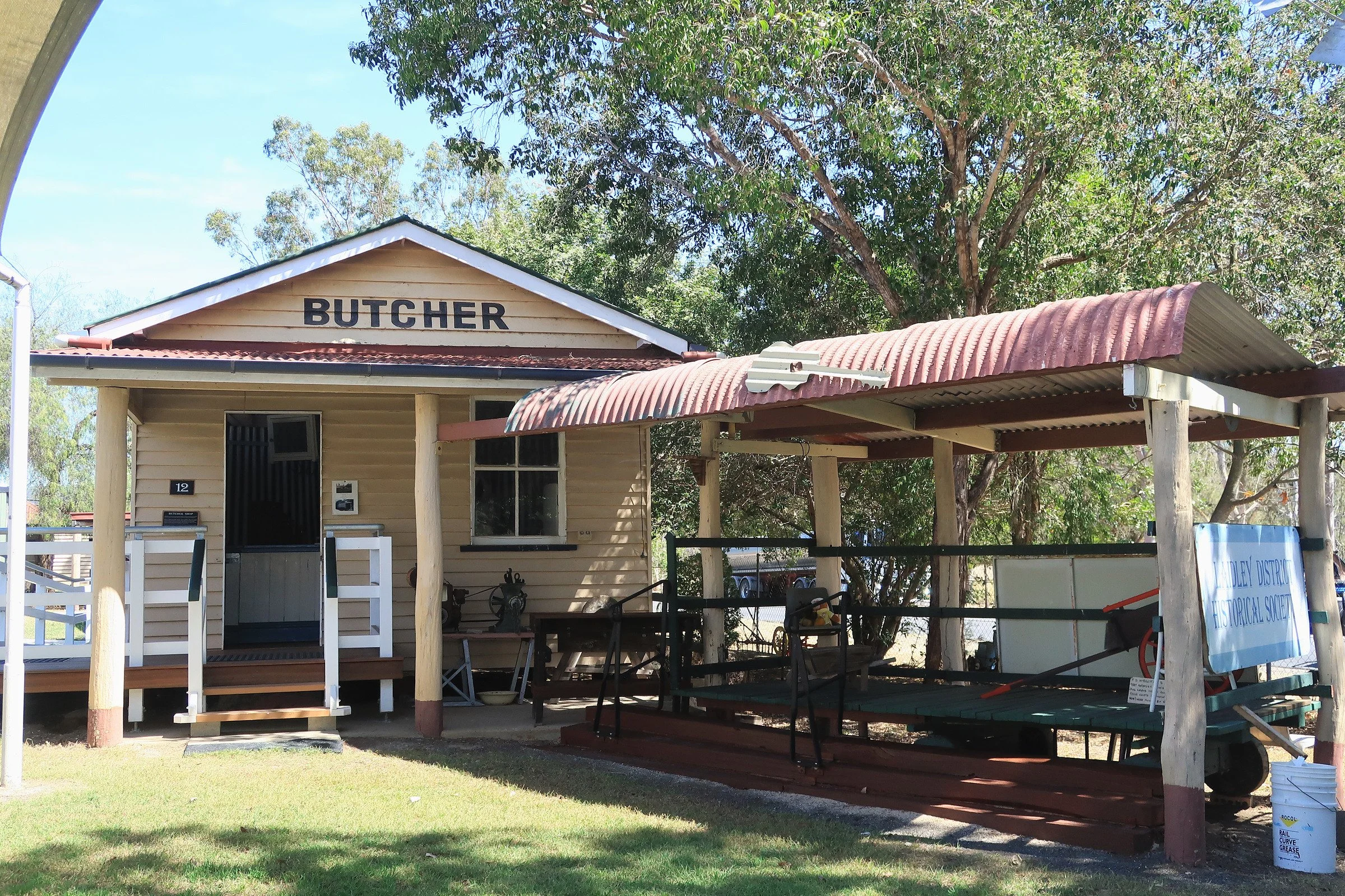 Small building with a sign that reads 'BUTCHER', and a front porch with steps, surrounded by trees and yard. There are various items, including chairs and barrels, in front of the building.