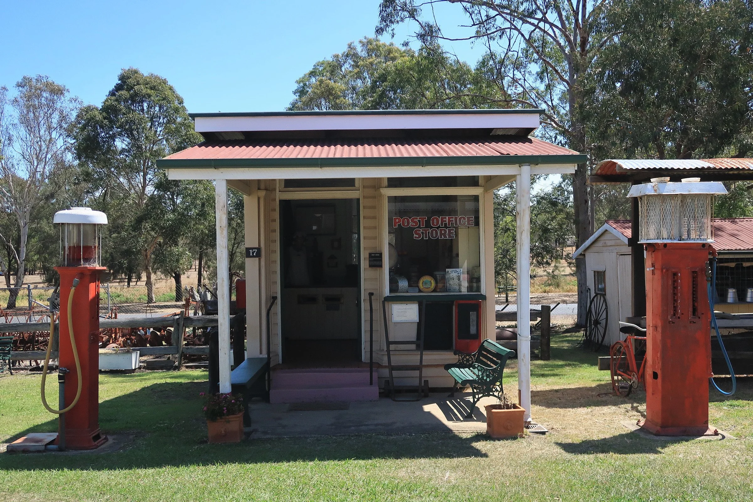 Small vintage post office store building with a sign in the window, surrounded by trees, and with vintage gas pumps nearby.