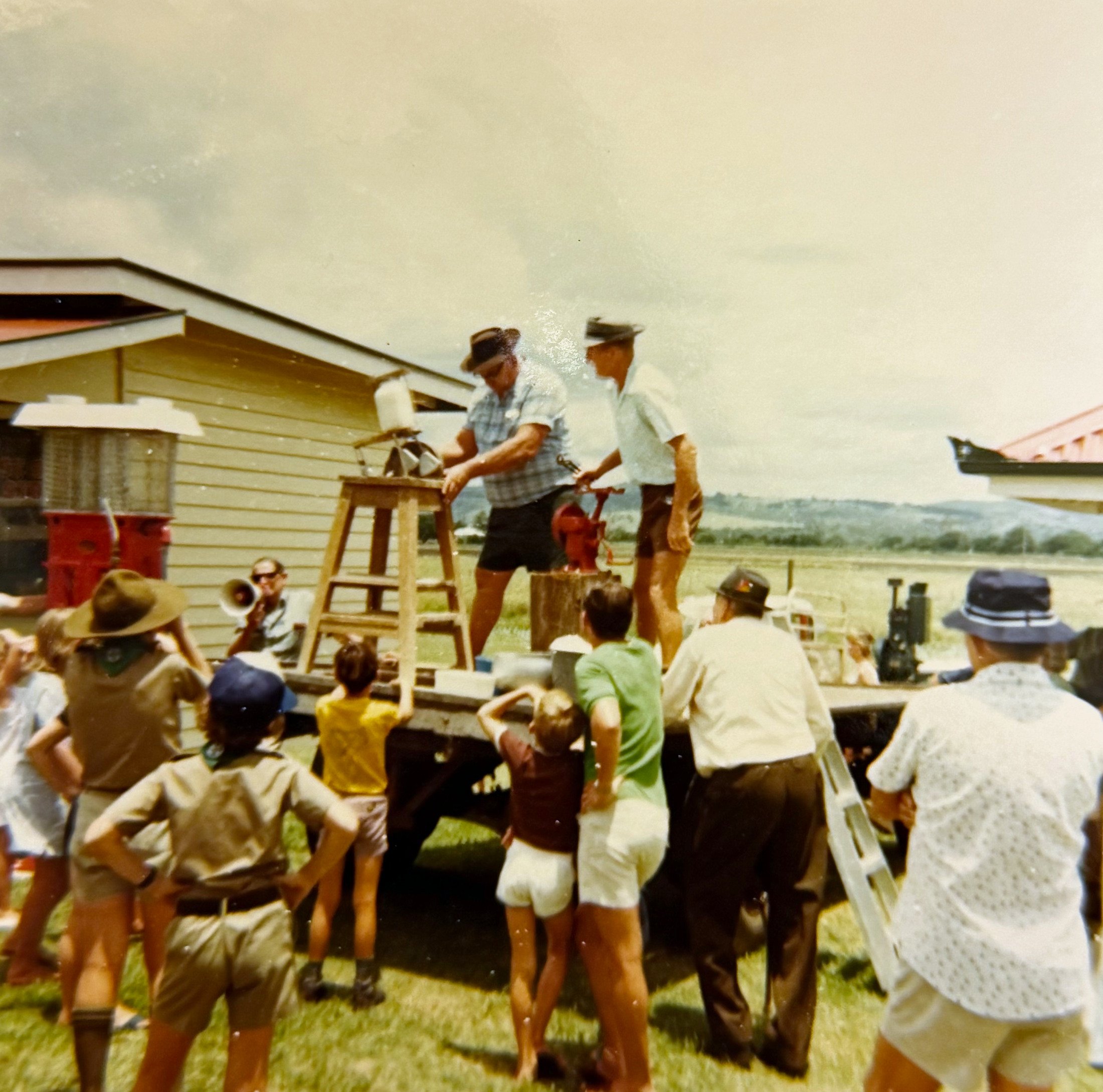 People gathered outdoors under a cloudy sky, sitting and standing near tables and a small wooden platform, possibly at a community event or gathering.
