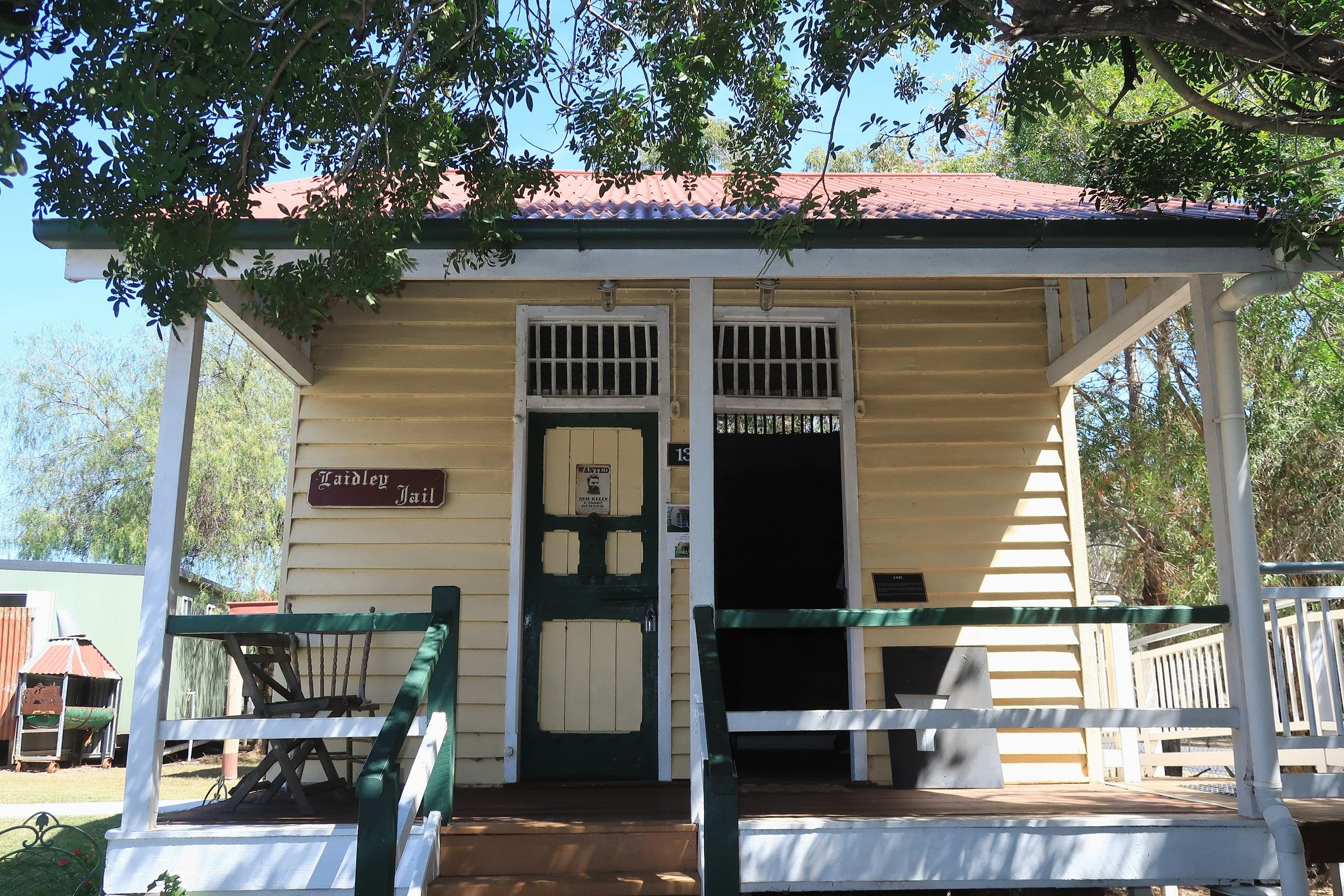 Front view of a yellow wooden house with a red tile roof, surrounded by trees and greenery, with signs indicating it is the Ladley Jail.
