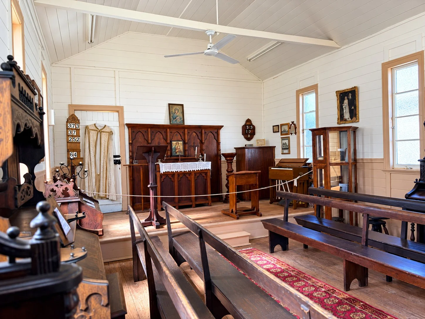 Interior of a small church with wooden pews, an altar with religious icons, candles, and a lace cloth, large windows with natural light, and religious artwork on the walls.