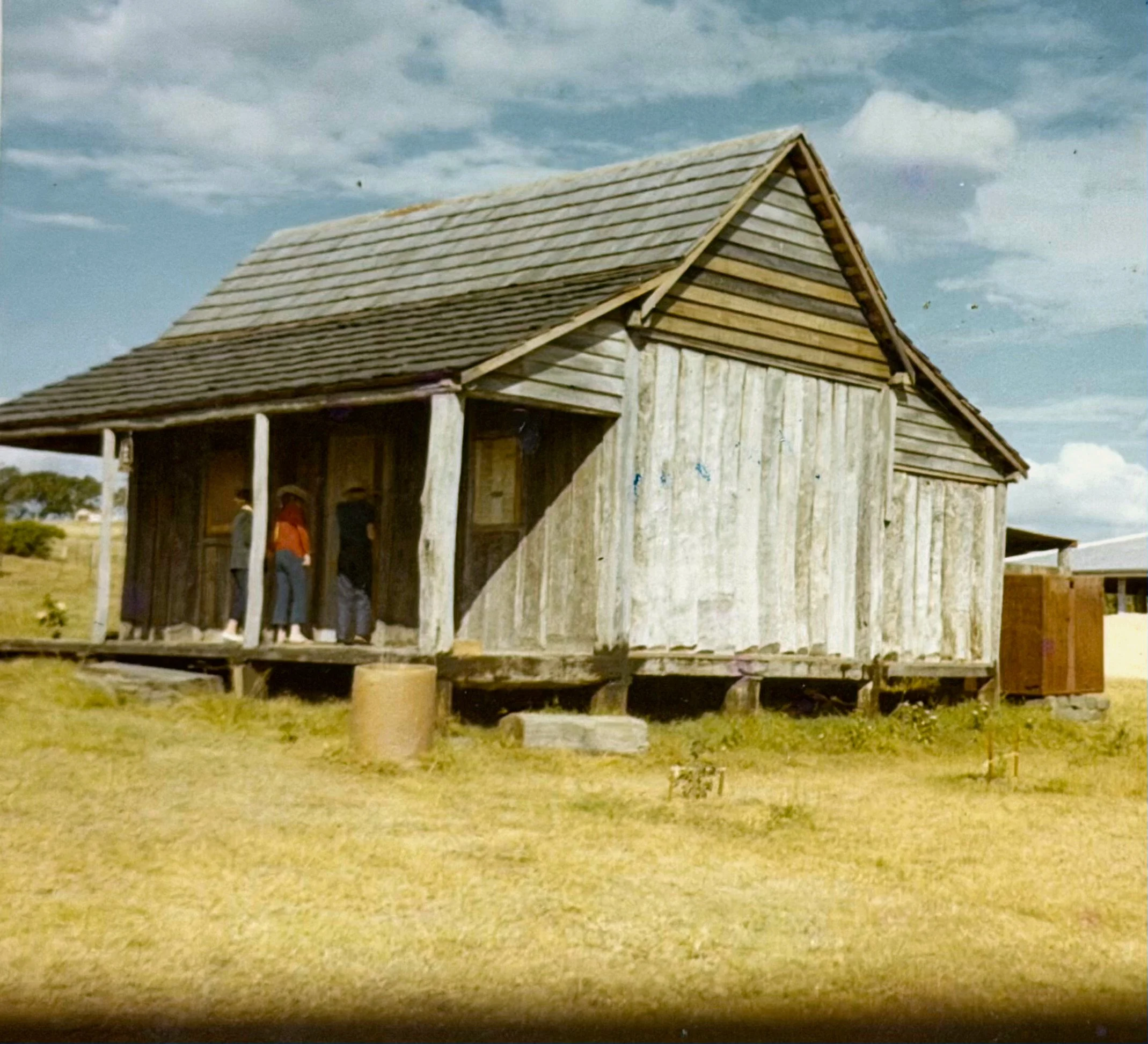 A rustic wooden barn with a sloped roof and weathered exterior, situated on a grassy field under a partly cloudy sky.
