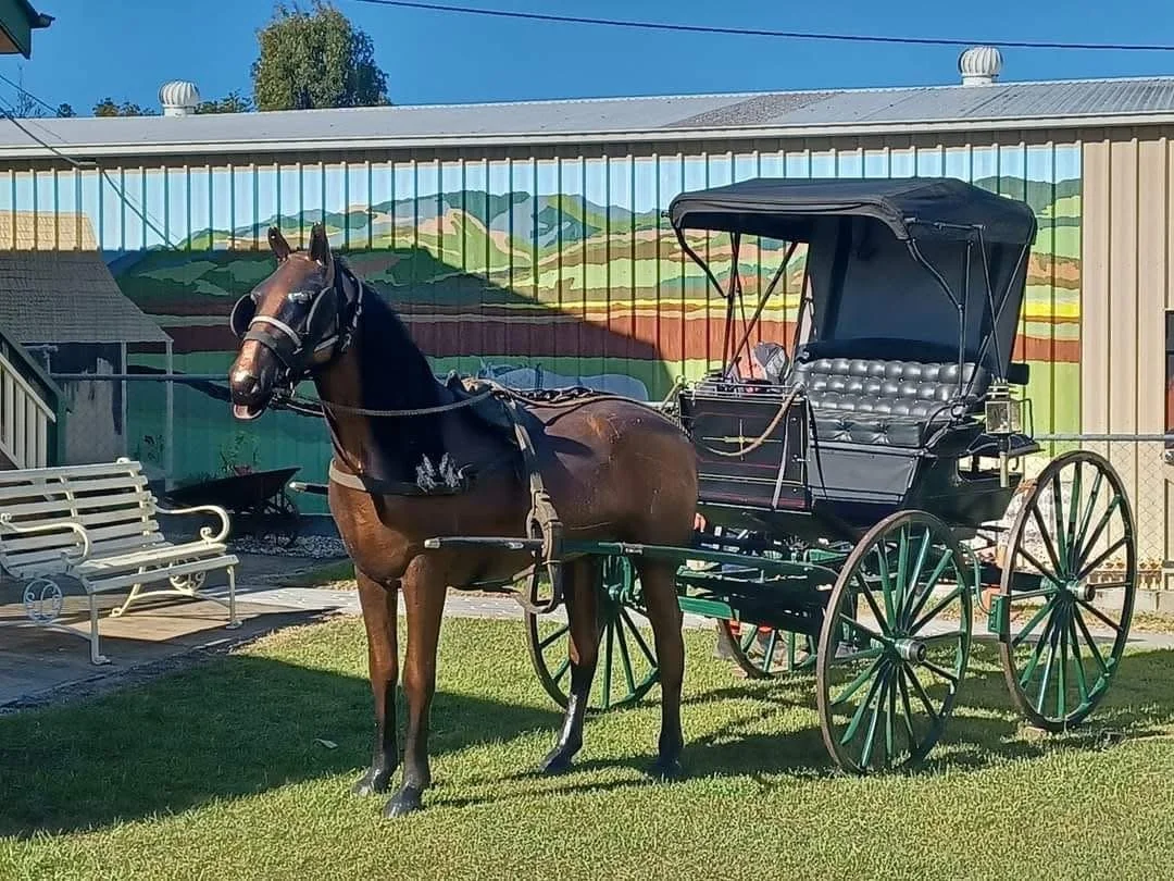 A brown horse hitched to a black horse-drawn carriage with black canopy, parked on grass in front of a colorful mural of rolling hills and blue sky.
