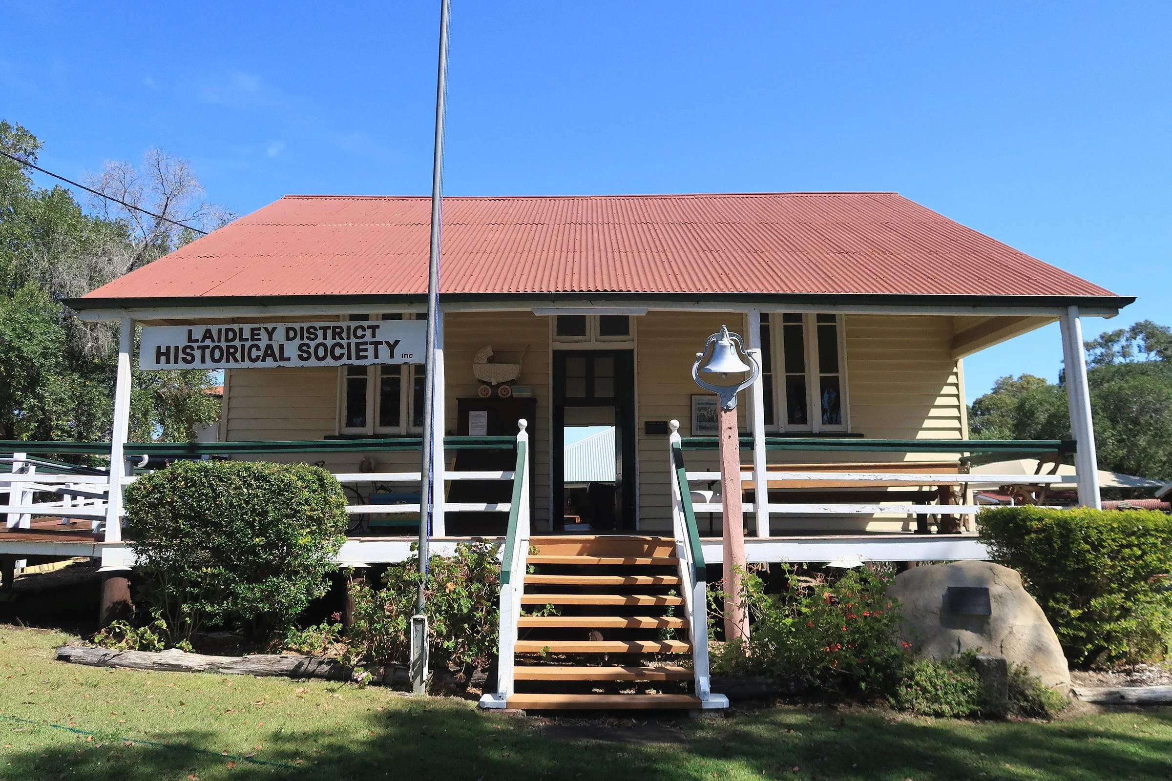 A yellow wooden building with a red metal roof, signs indicating it is part of the Laidley District Historical Society, and a bell mounted in front. There are stairs leading up to the porch and surrounding greenery, trees, and a clear blue sky.