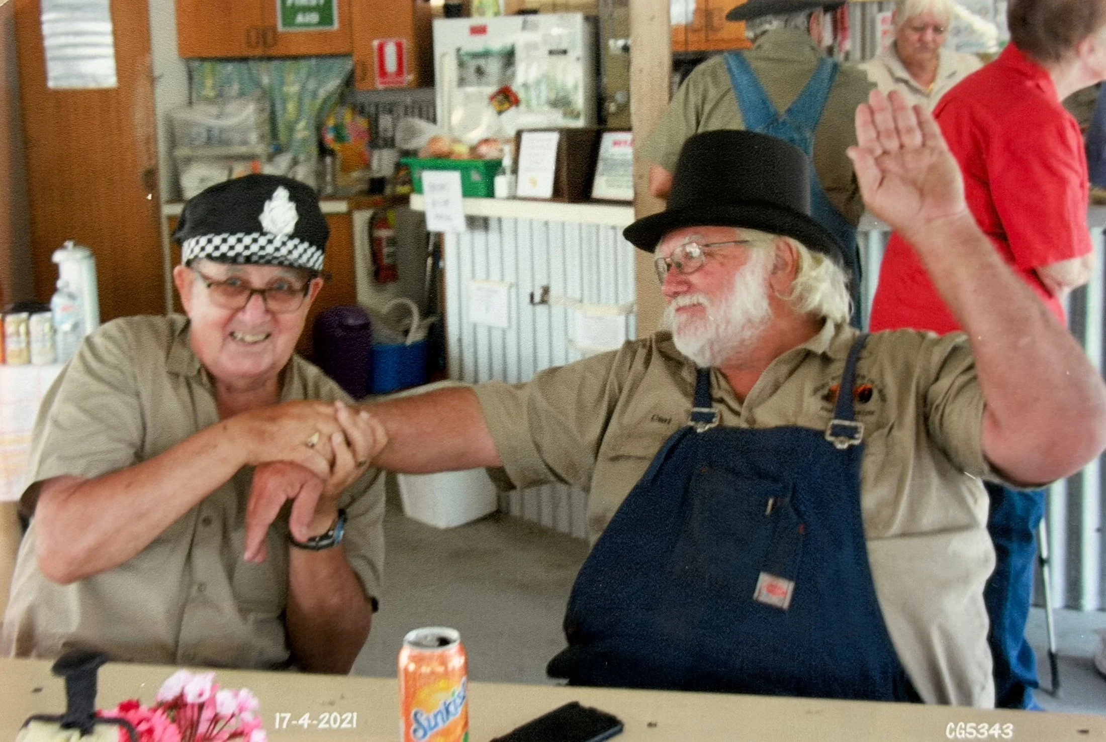 Two older men, one with glasses and a black and white checkered hat, and the other with a black hat and gray beard, are arm wrestling and smiling in a store or community center. There are others in the background, and a table with snacks and a vase with pink flowers in the foreground.