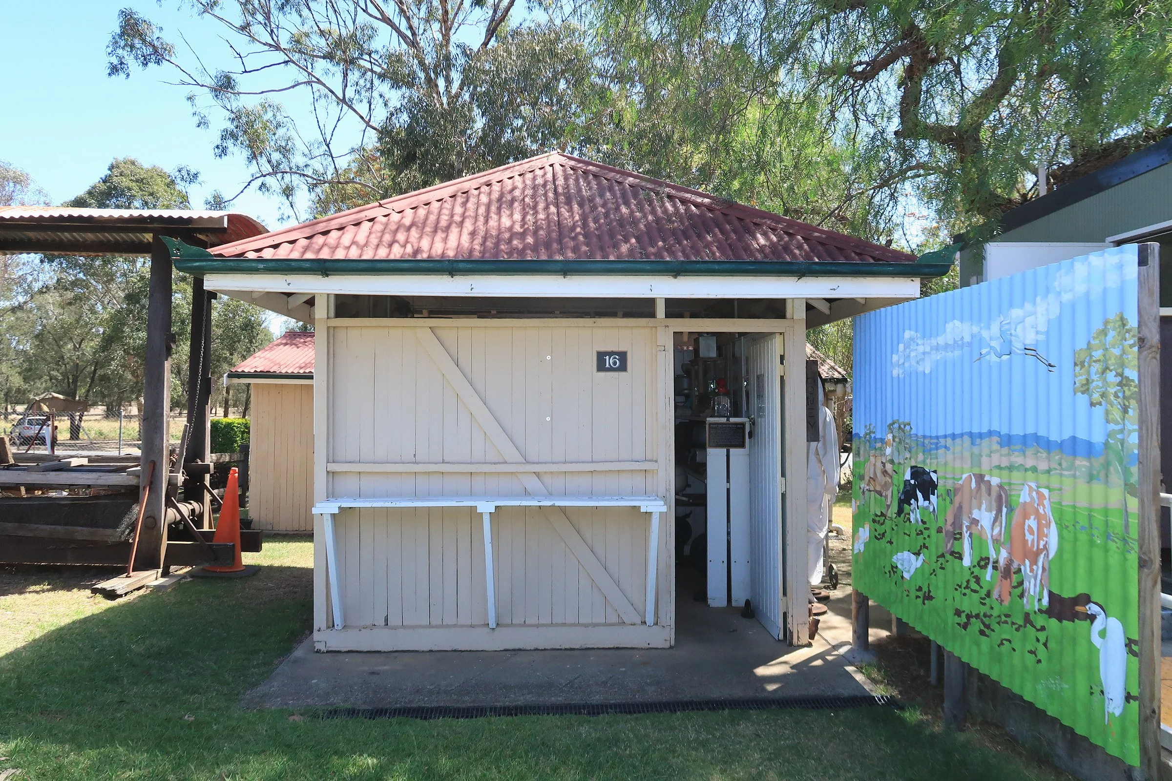 A small white wooden shed with a red tile roof, a bench outside, and a colorful outdoor curtain or banner to the right. There are trees and a clear blue sky in the background.