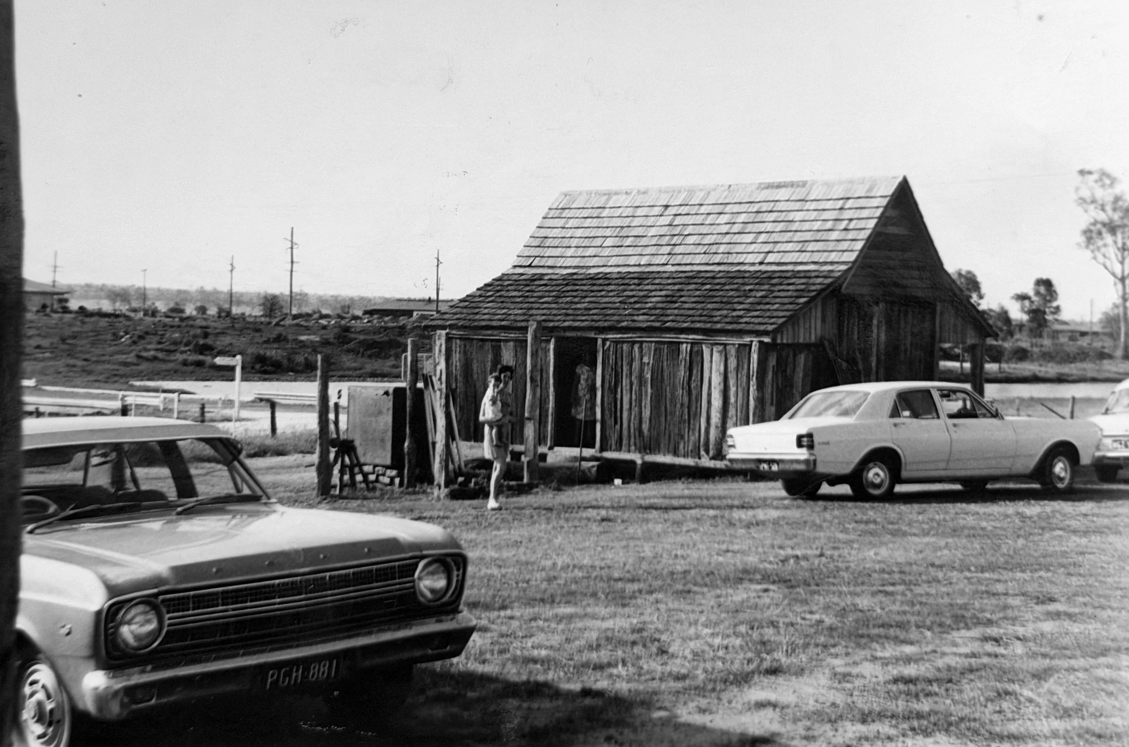 Black and white photo of an old wooden barn with a tiled roof, two men standing in front, a few cars parked nearby, utility poles in the background, and trees in the distance under a clear sky.
