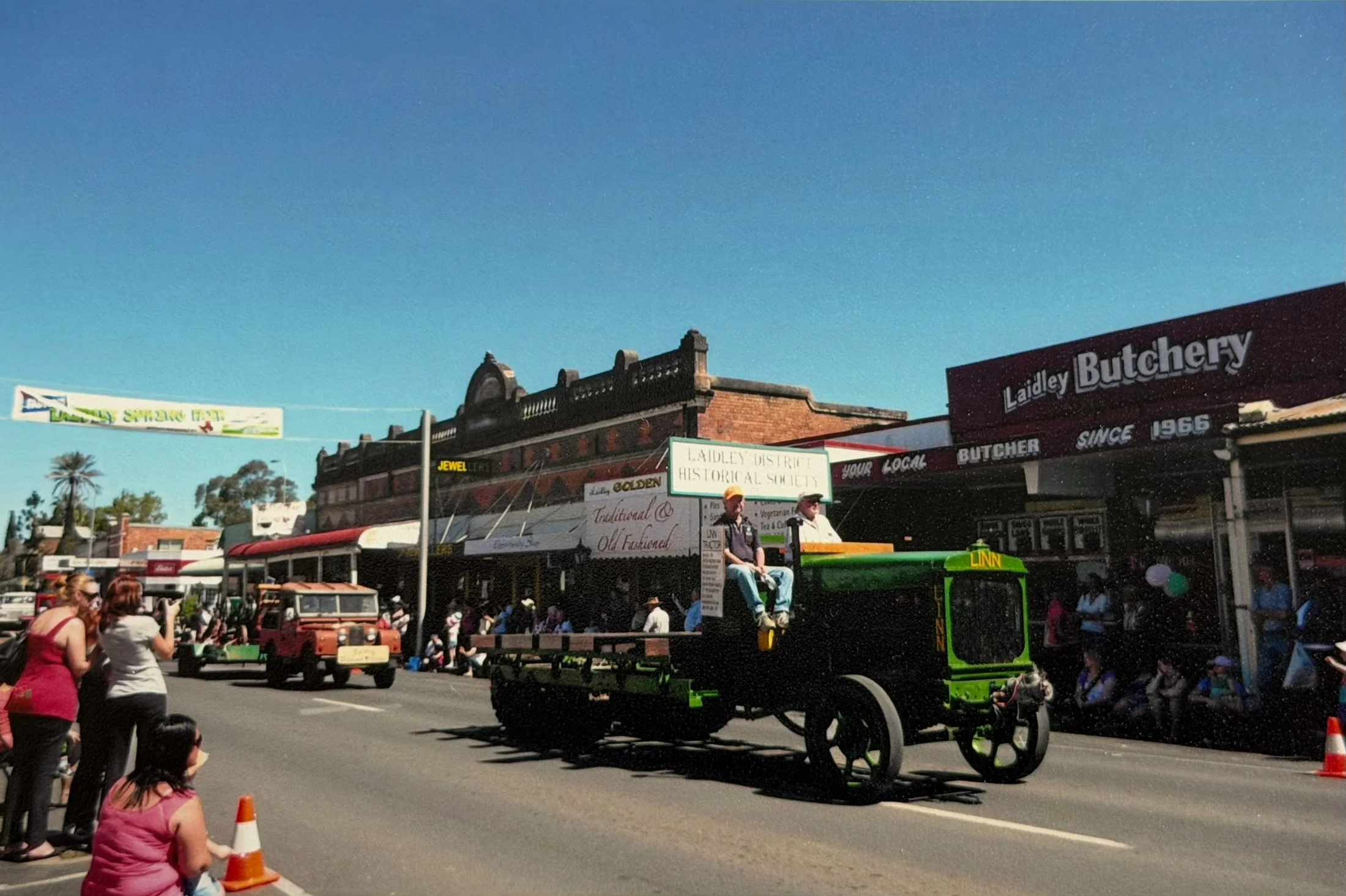Street scene with people waiting, a horse-drawn carriage in the foreground, historic brick buildings, and signs indicating a local district and historic safety event.