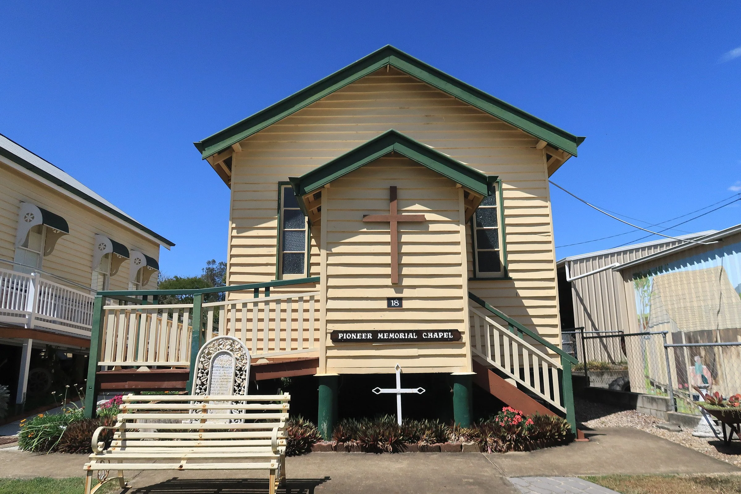 A small wooden church with a cross on the front, stairs leading up to it, and the sign 'Pioneer Memorial Chapel' on the facade. There are benches, flowers, and other structures around in a sunny outdoor setting.
