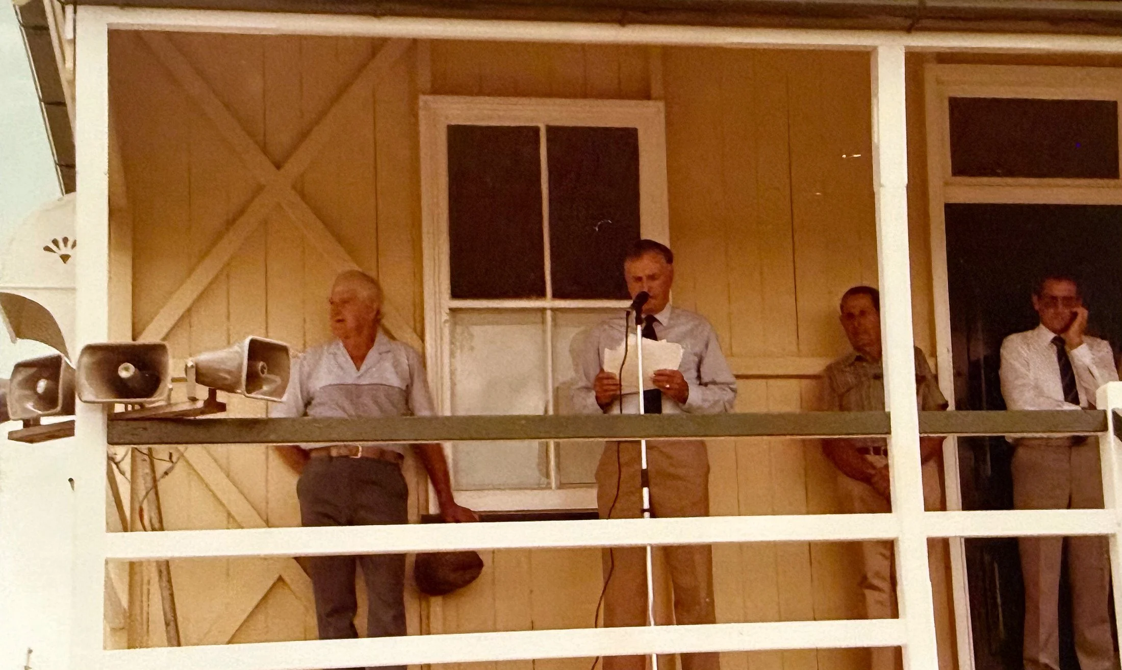 Four people stand on a wooden balcony with a tan wall background. One woman is speaking into a microphone, holding papers, while three men stand nearby, listening or waiting.