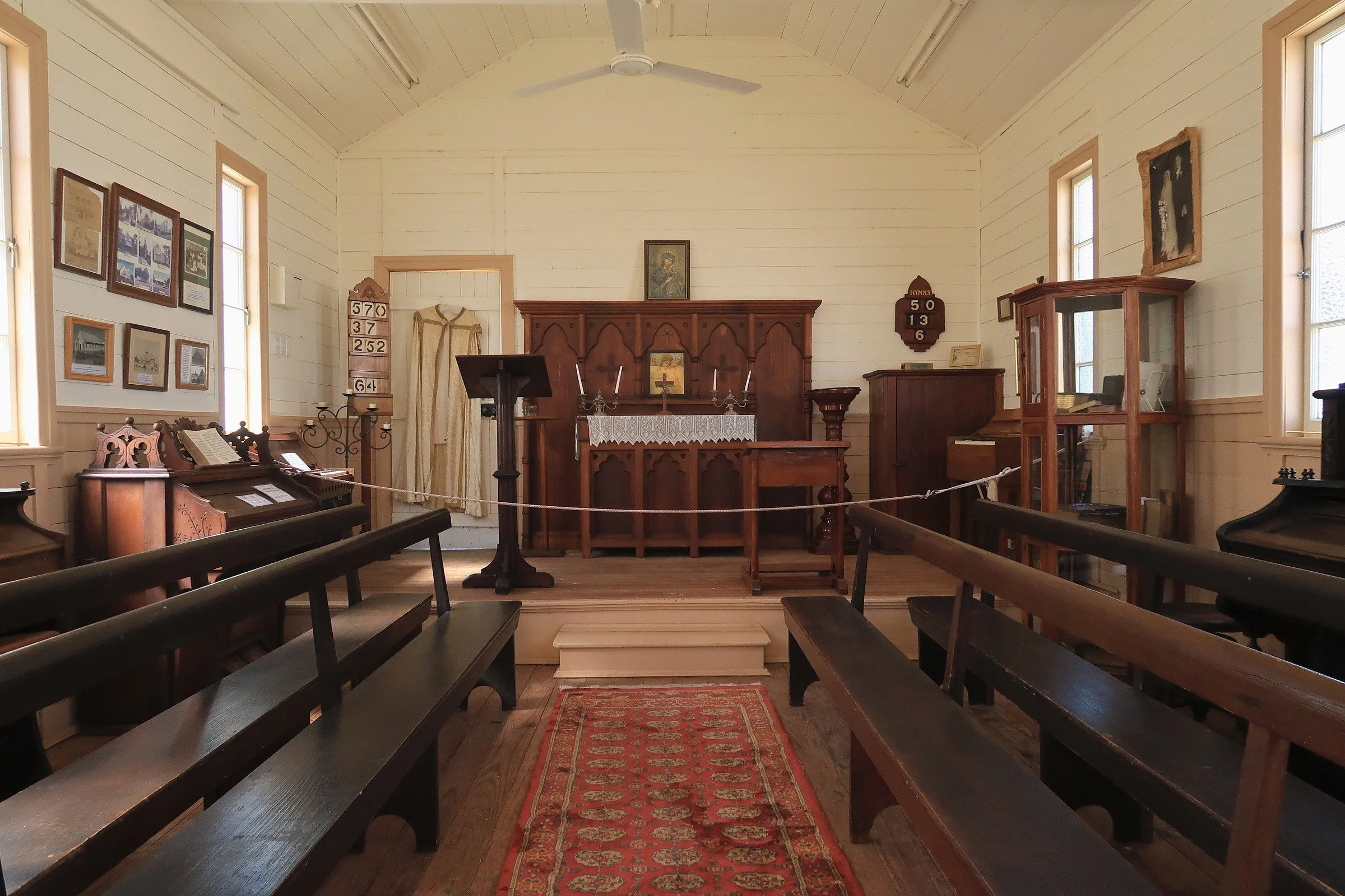 Interior of a small church with wooden pews, a red carpet runner, a wooden altar, framed religious and historical pictures on the walls, and windows letting in natural light.