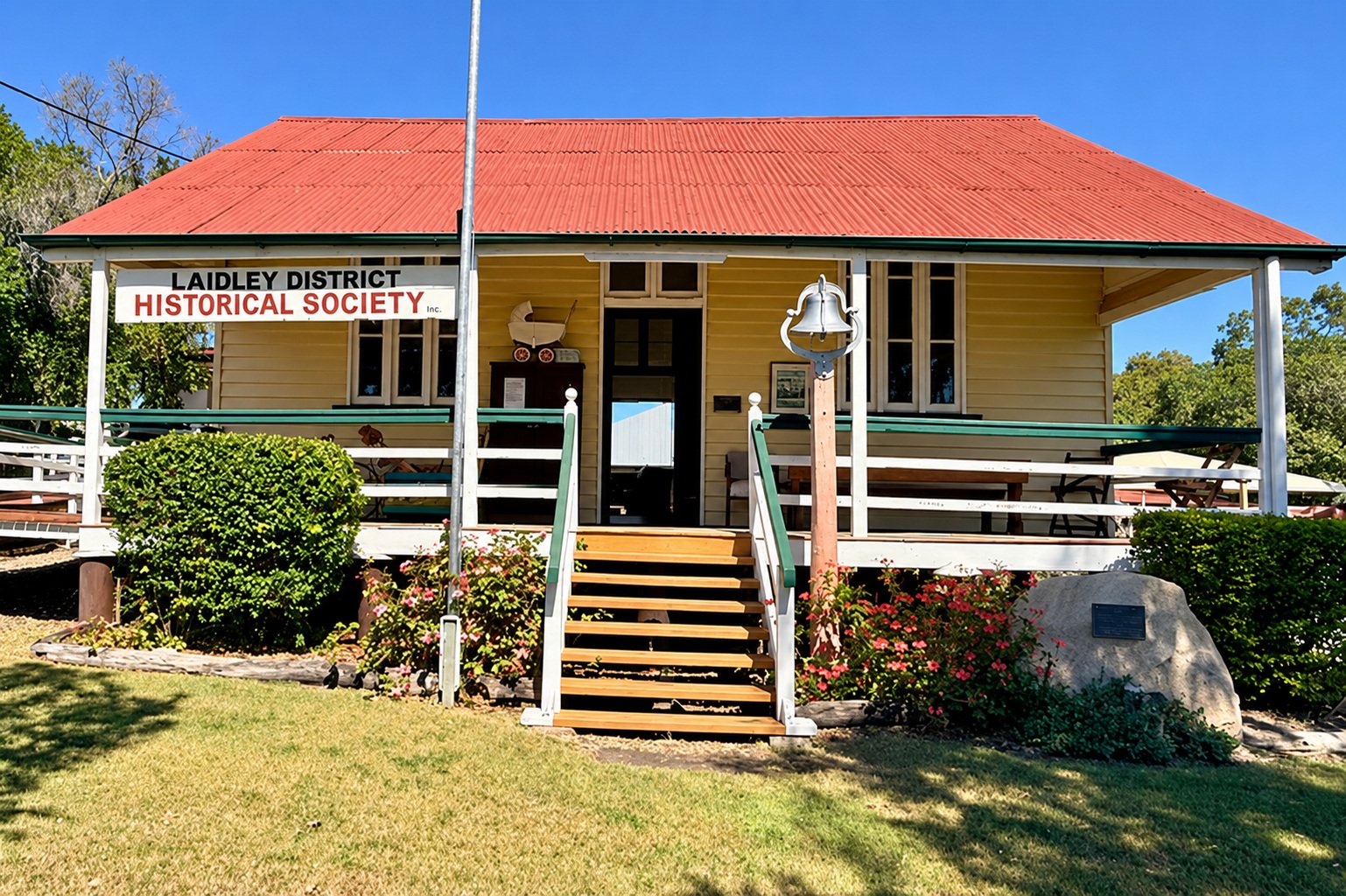 A yellow wooden building with a red metal roof, labeled 'Ladley District Historical Society Inc.' with a porch, wooden stairs, and garden flowers in front, under a blue sky.