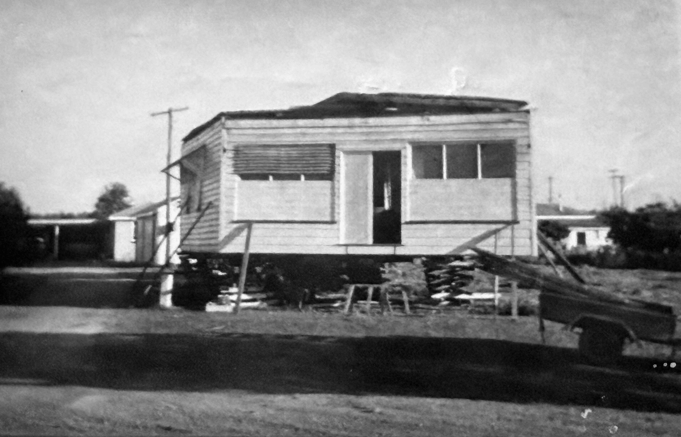 Black and white photo of a house under construction with unfinished walls and construction materials around, set in a rural area with utility poles in the background.
