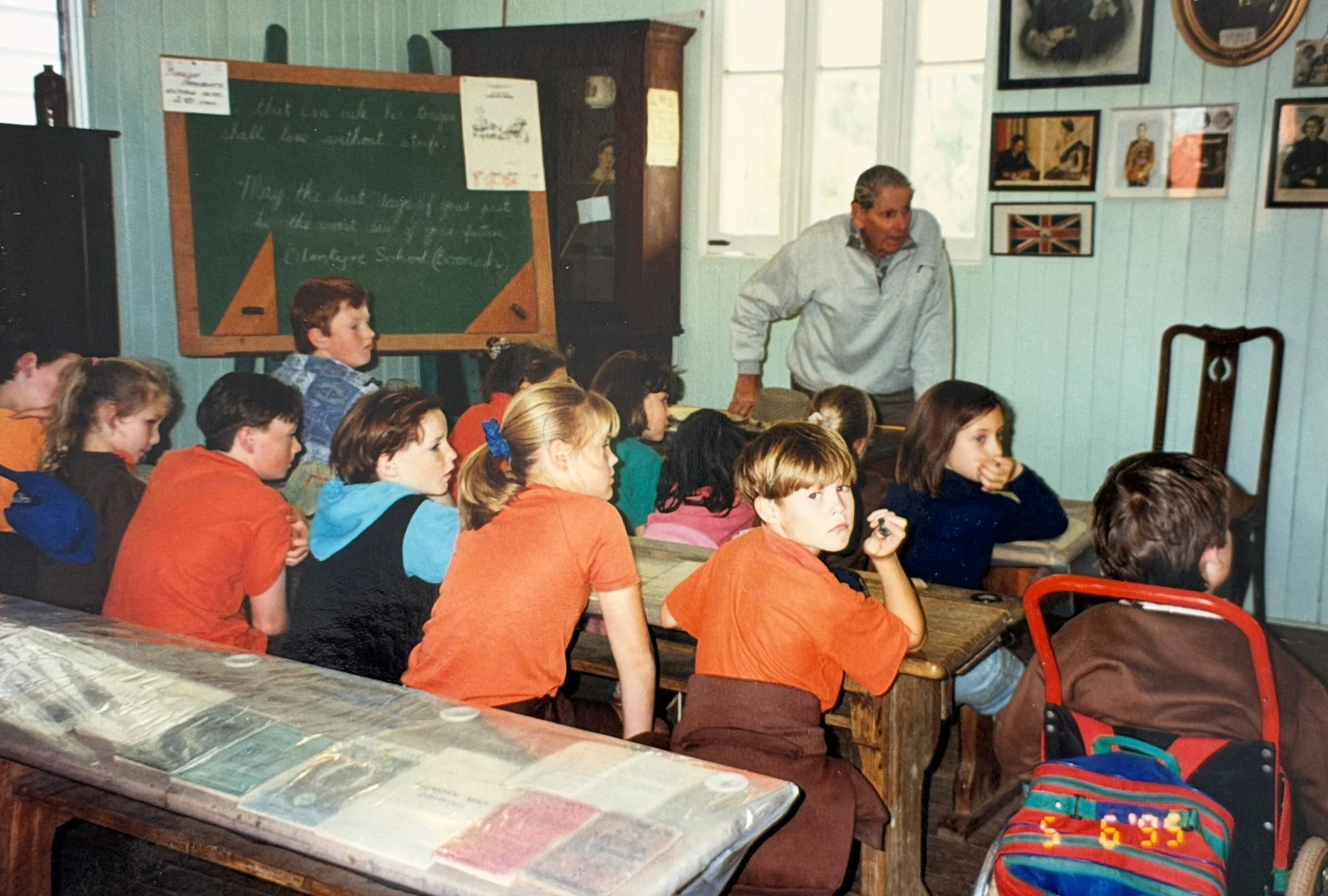 A classroom with children sitting at desks, paying attention to a teacher standing next to a chalkboard. The classroom has framed pictures on the wall, including a British flag, and windows letting in natural light.