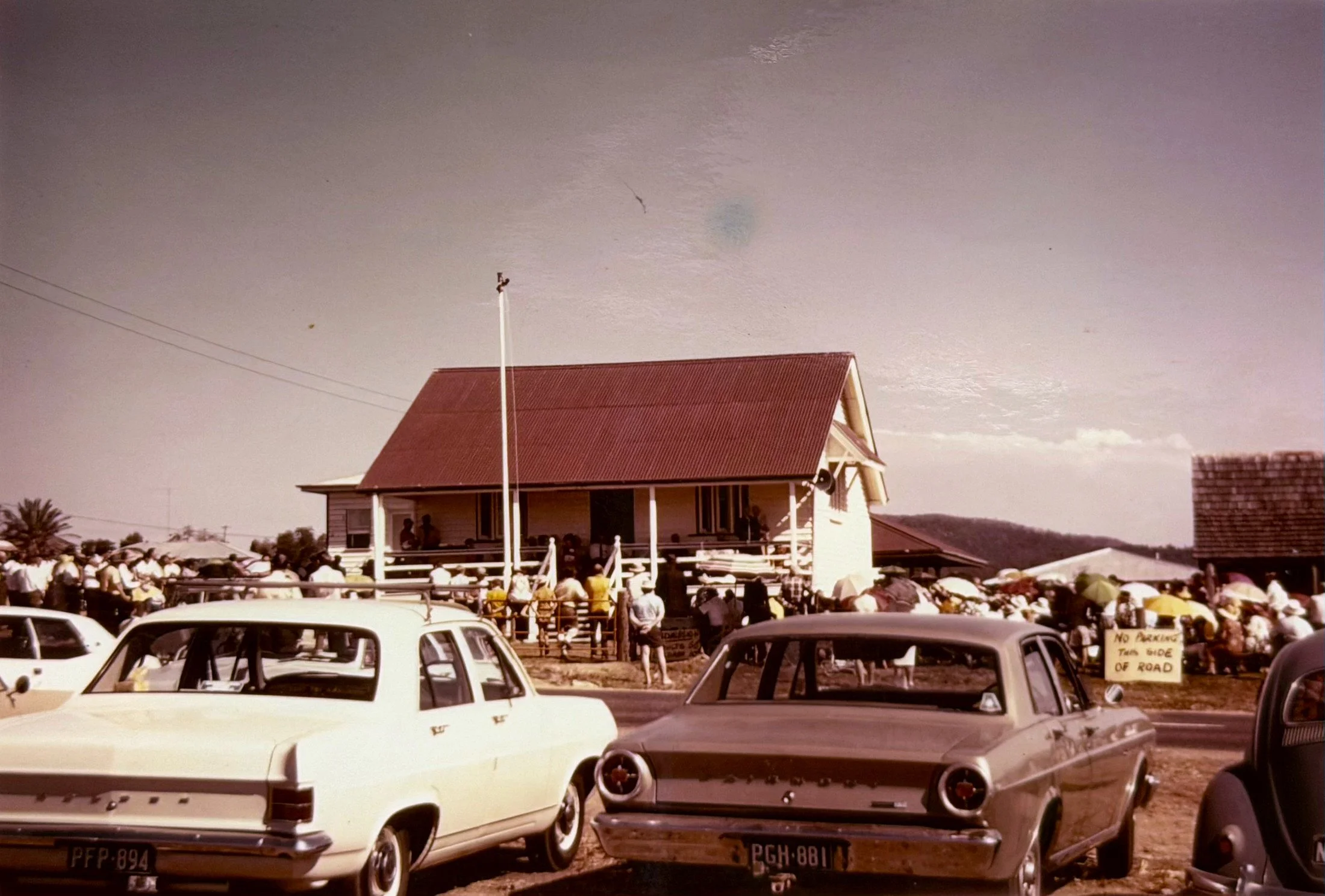 A crowd gathered at an outdoor event in front of a house with people seated on the porch. Cars are parked in the foreground, and a sign on one car reads 'No parking, ten side of road.' The sky is mostly clear with a few clouds.