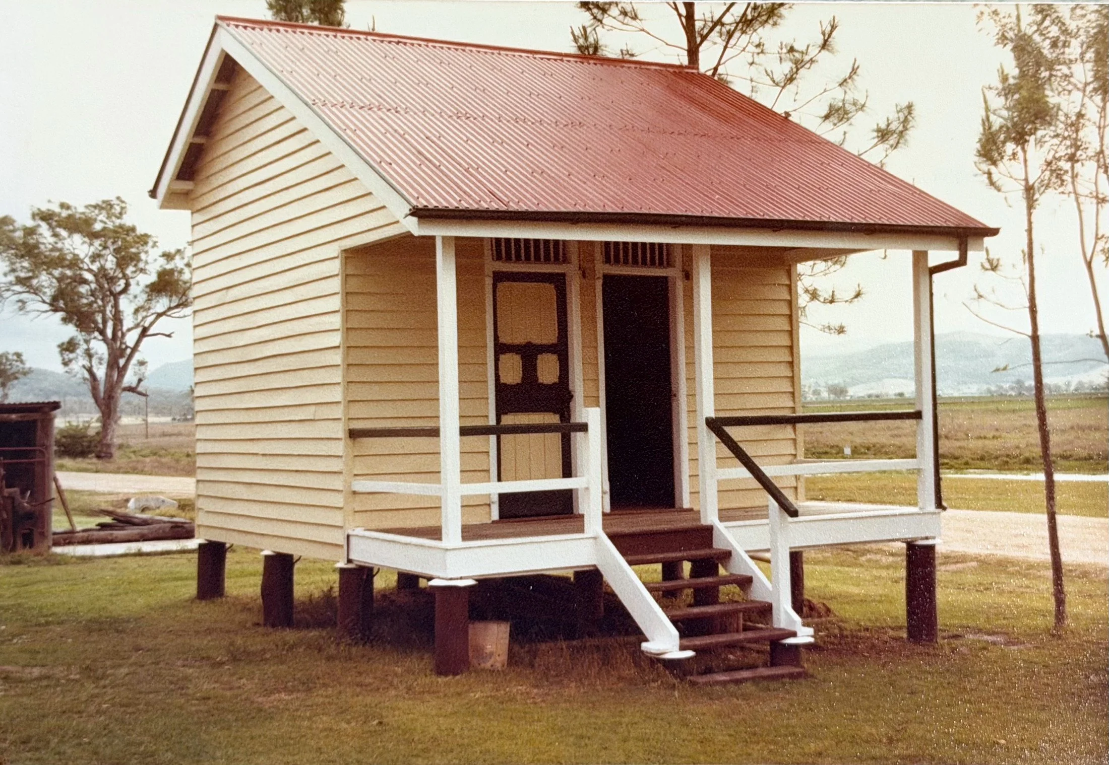 A small tan wooden house with a red corrugated metal roof, stairs leading to a front porch, and surrounded by trees and open land.