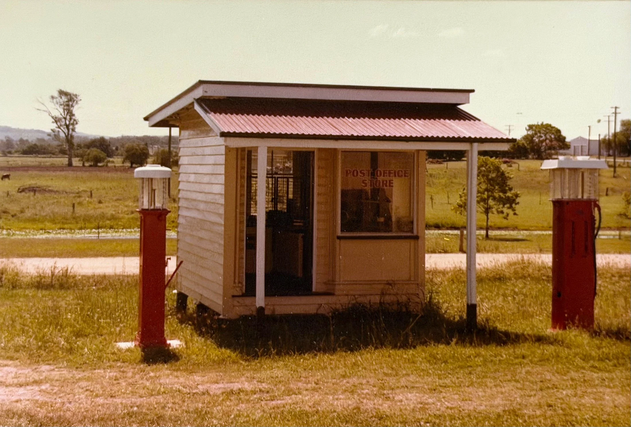 A small wooden post office store with a slanted red metal roof, situated in a rural landscape with open fields, trees, and utility poles in the background.