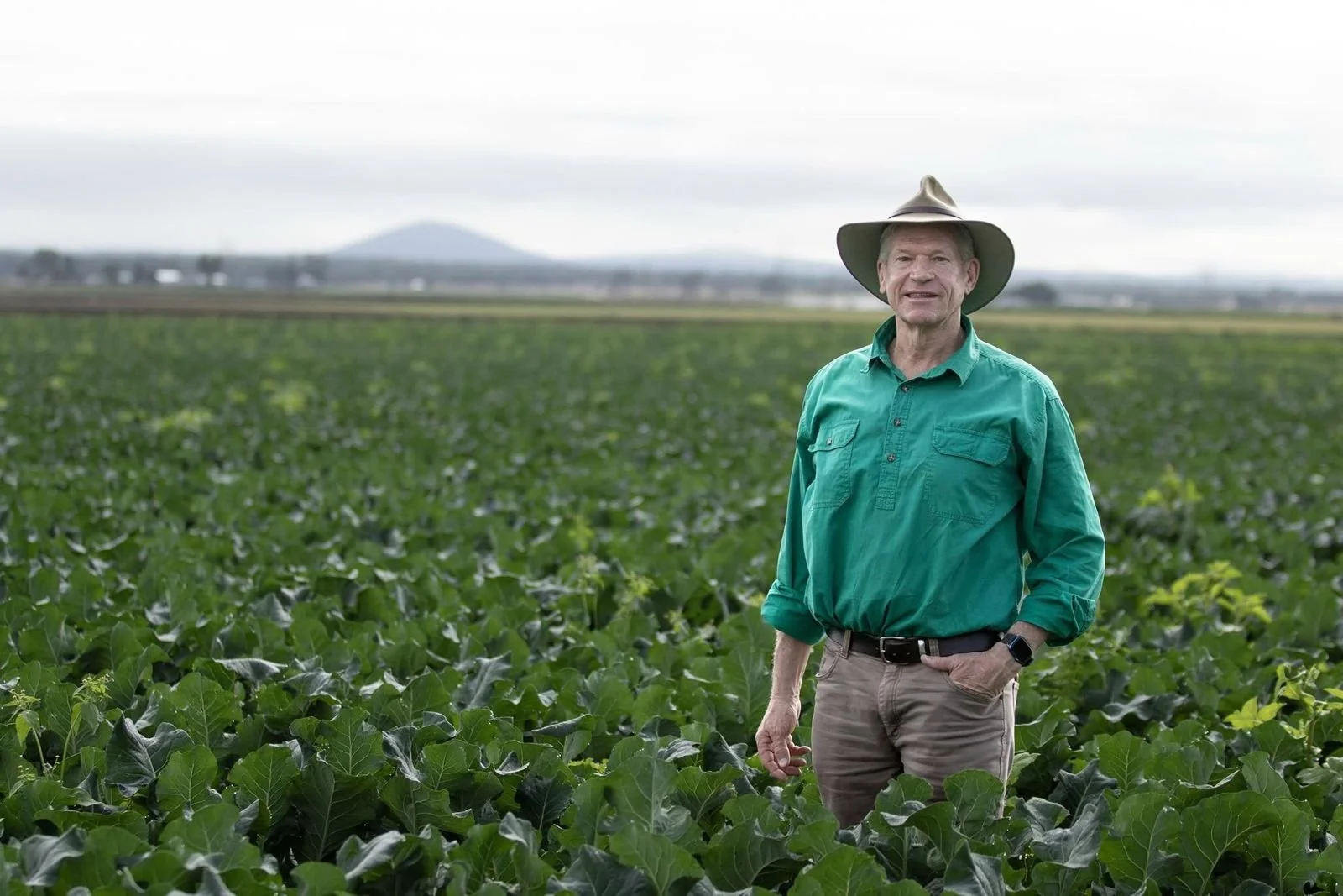 A man in a teal shirt and gray pants standing in a lush green farm field with a wide-brimmed hat, overcast sky, and distant landscape in the background.