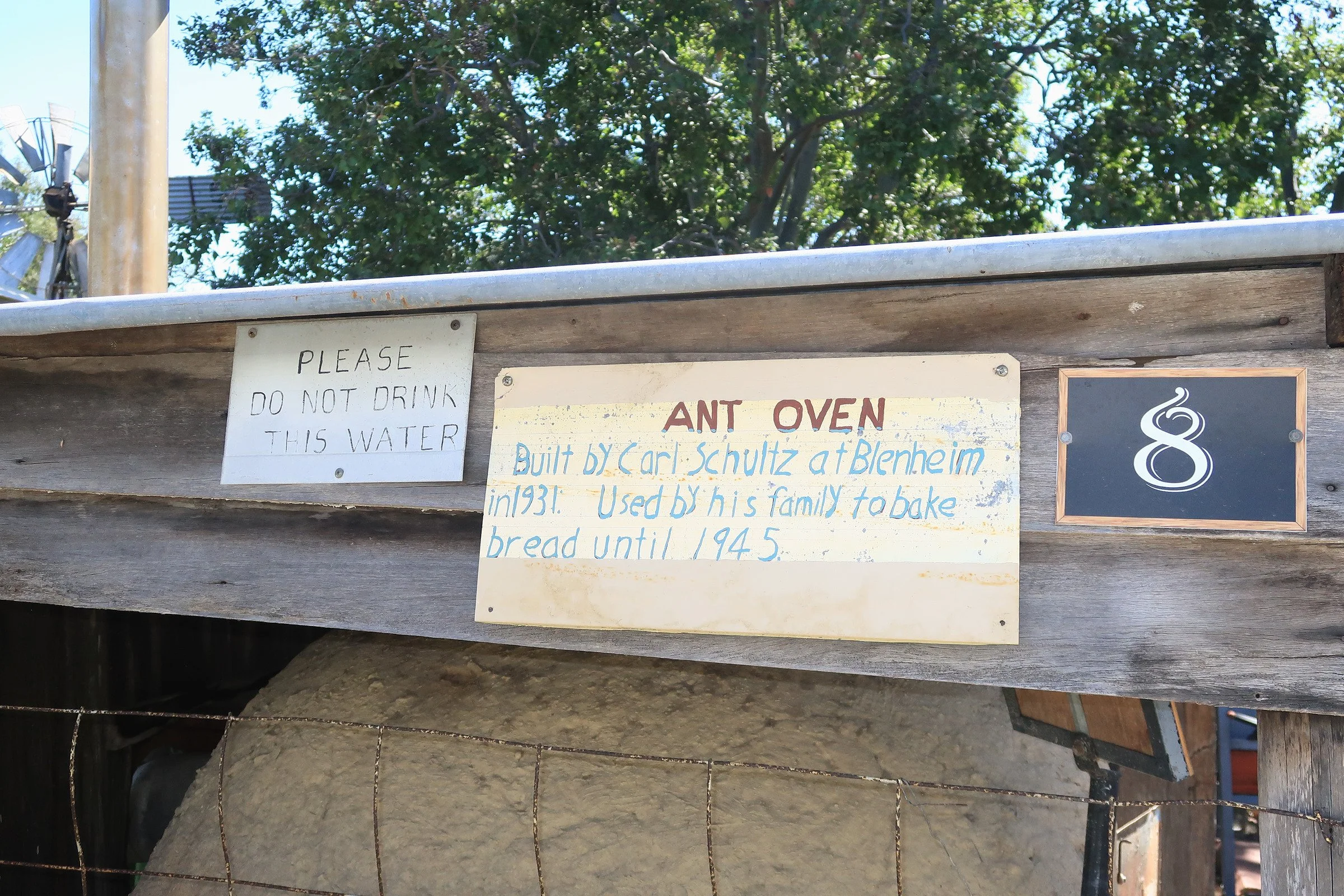 Photograph of a wooden and metal structure with handwritten signs mentioning an ant oven, a request not to drink the water, and historical information about its builder Carl Schultz.