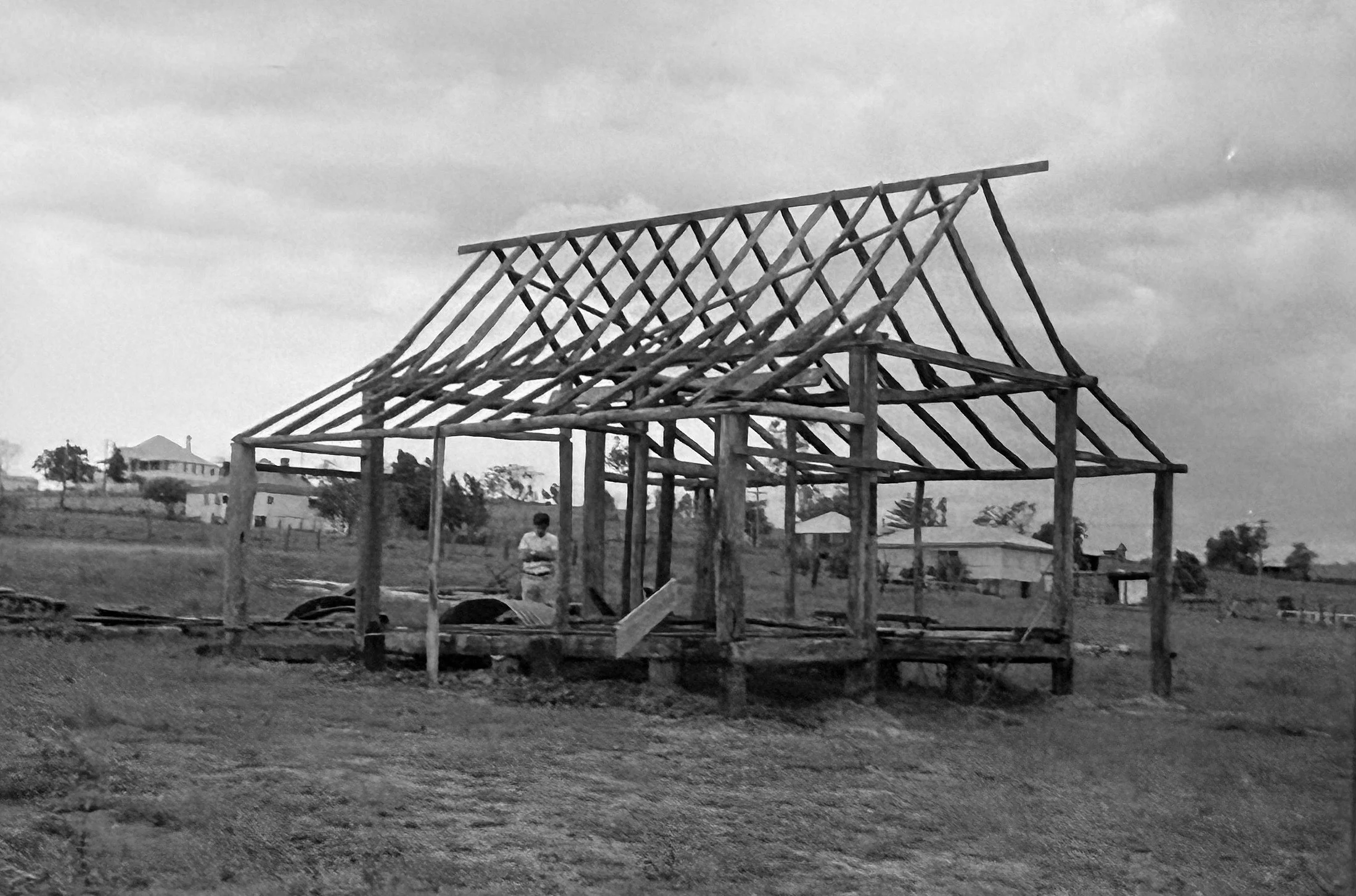 A wooden house frame under construction with a peaked roof, set in an open field with distant buildings and trees in the background.