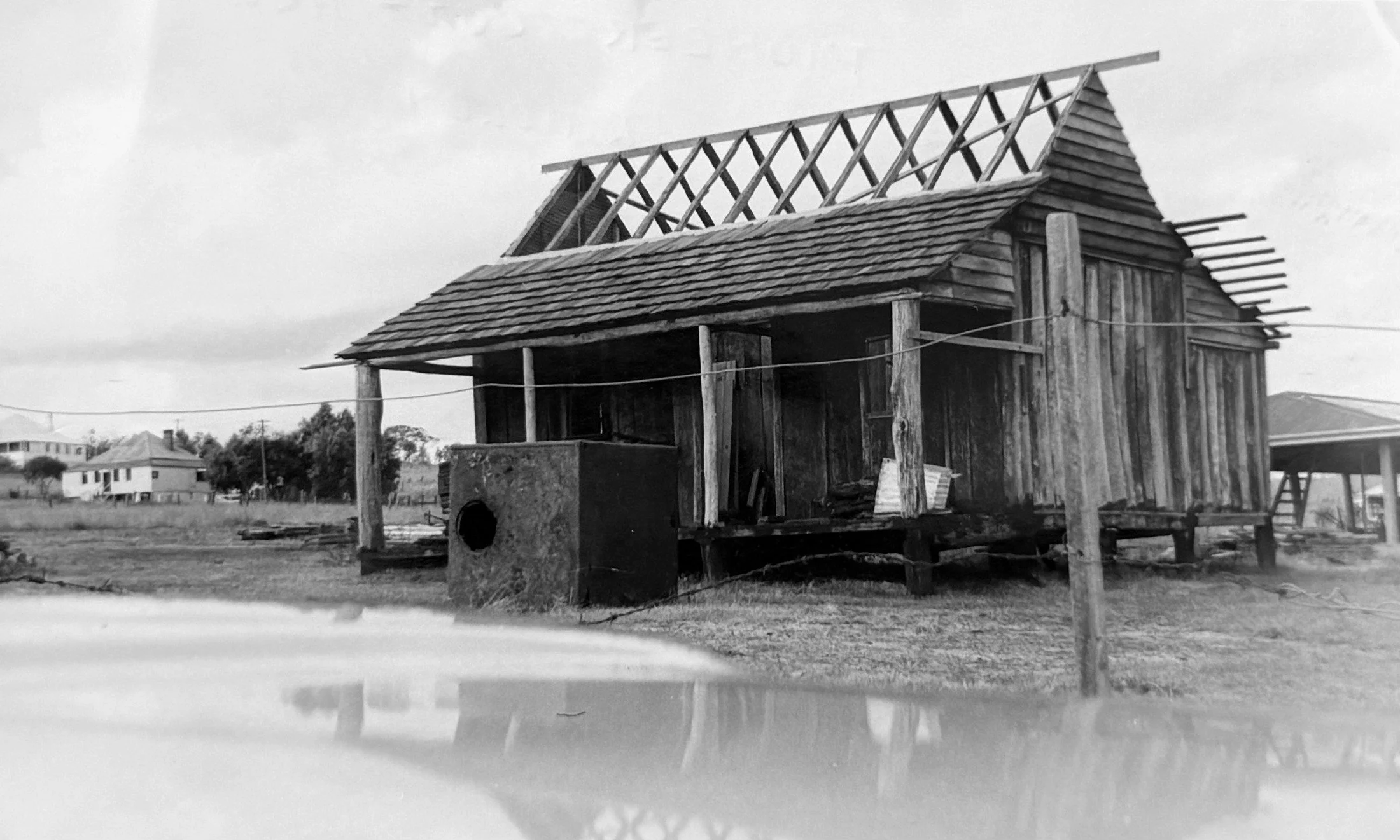 A black and white photo of an unfinished wooden house on stilts, with a roof partially built, in a rural area with other houses in the background.