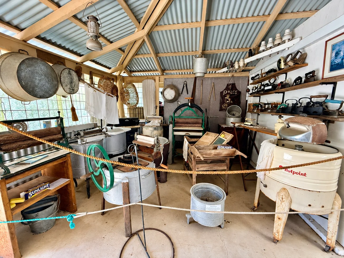A vintage laundry room or workshop filled with old-fashioned washing machines, irons, and other antique household items on shelves and surfaces, with a rustic wooden ceiling and large windows.