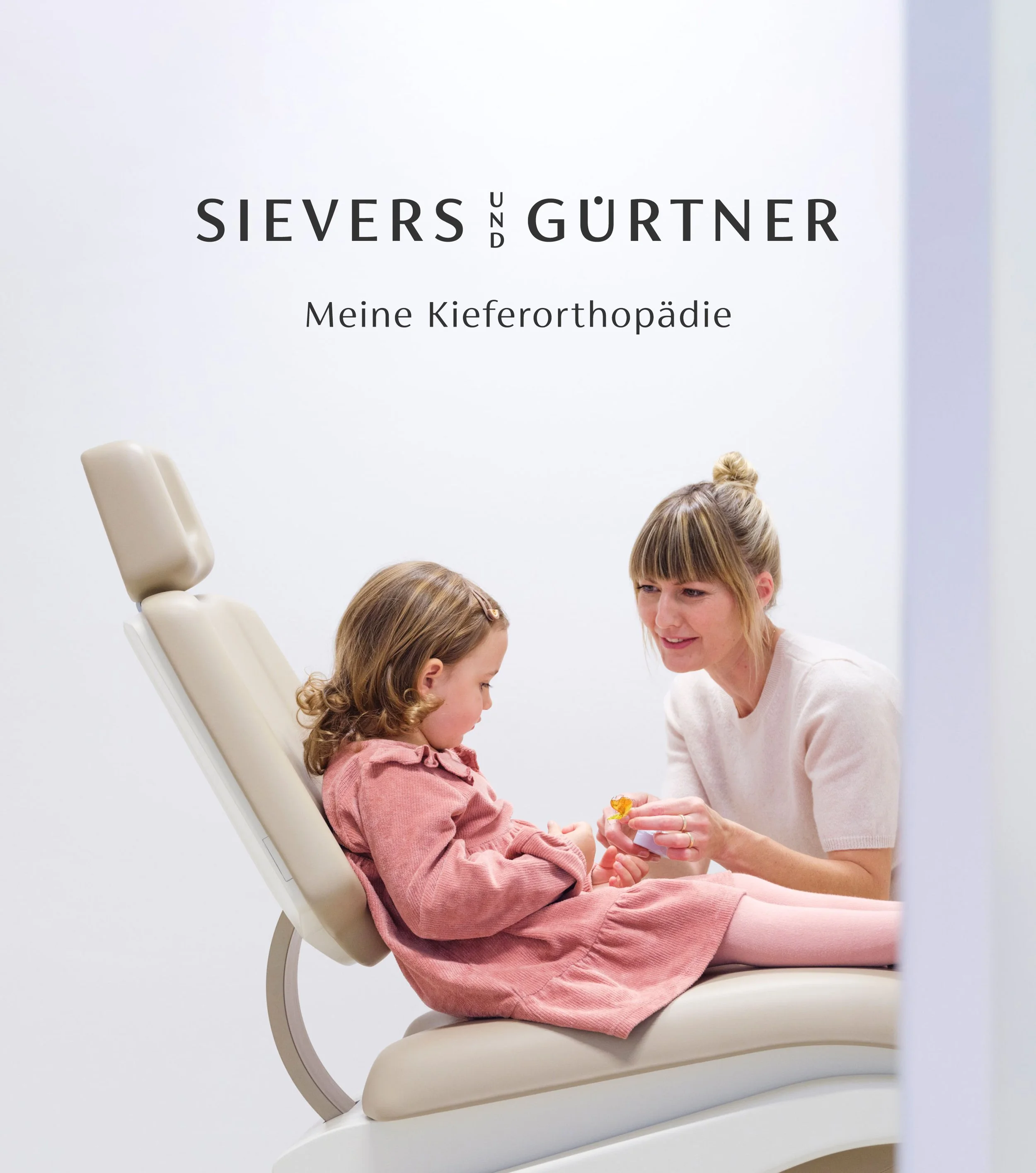 Eine junge Frau berät ein Mädchen mit braunen lockigen Haaren, die auf einem Therapiesessel sitzt, in einem hellen Raum in einer kieferorthopädischen Praxis. Die junge Frau trägt ein weißes Oberteil und wirkt freundlich.