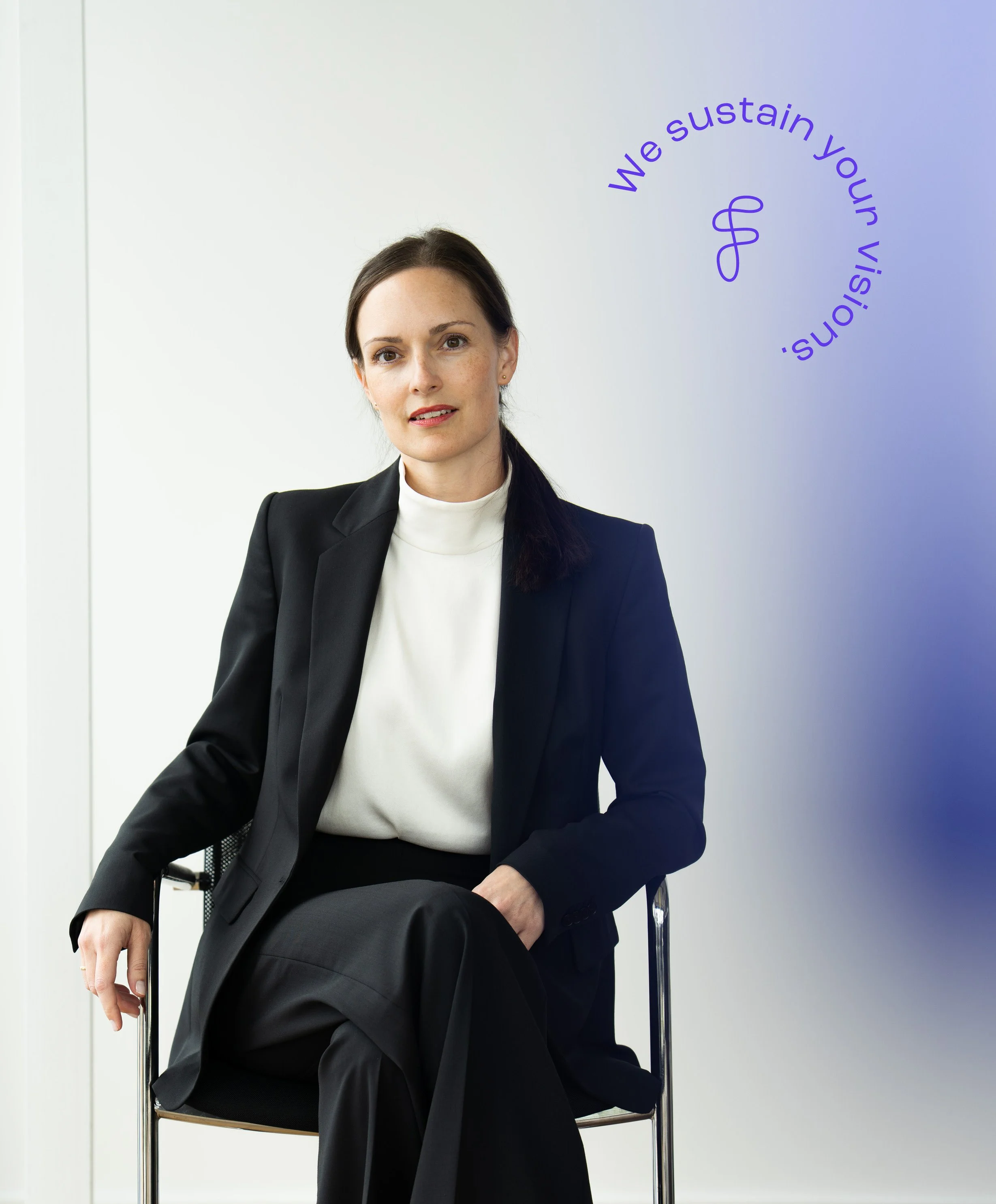 Eine Frau sitzt auf einem Stuhl und trägt einen schwarzen Anzug mit einem weißen Oberteil. Im Hintergrund ist eine weiße Wand, und an der Wand schwingt ein dekorativer Schriftzug mit dem Satz "We sustain your visions", der in einem Bogen angeordnet ist.