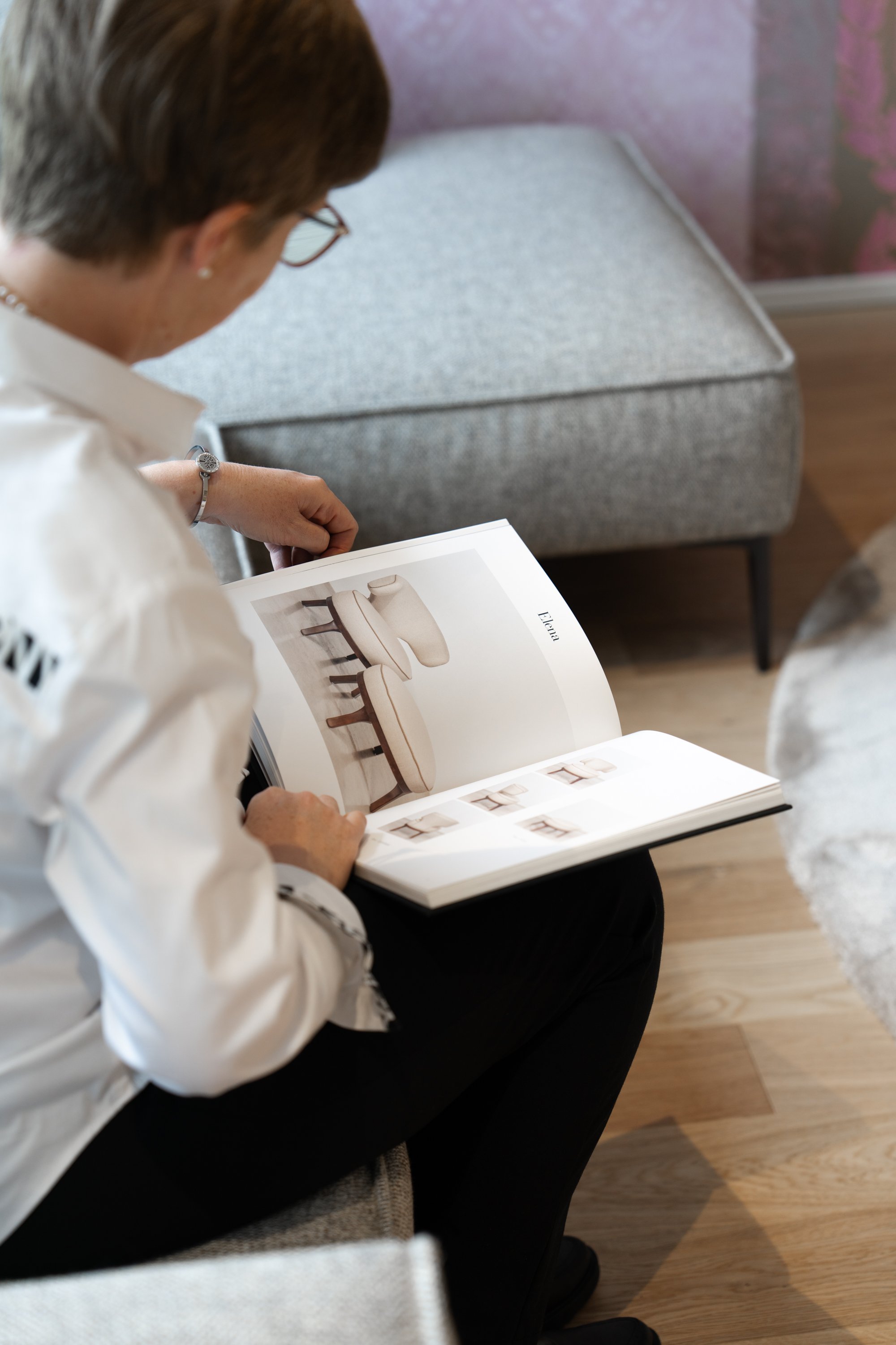 Frau liest in einem Katalog mit Möbeln, sitzt auf einem Stuhl vor einem grauen Sofa in einem Raum mit Holzboden.